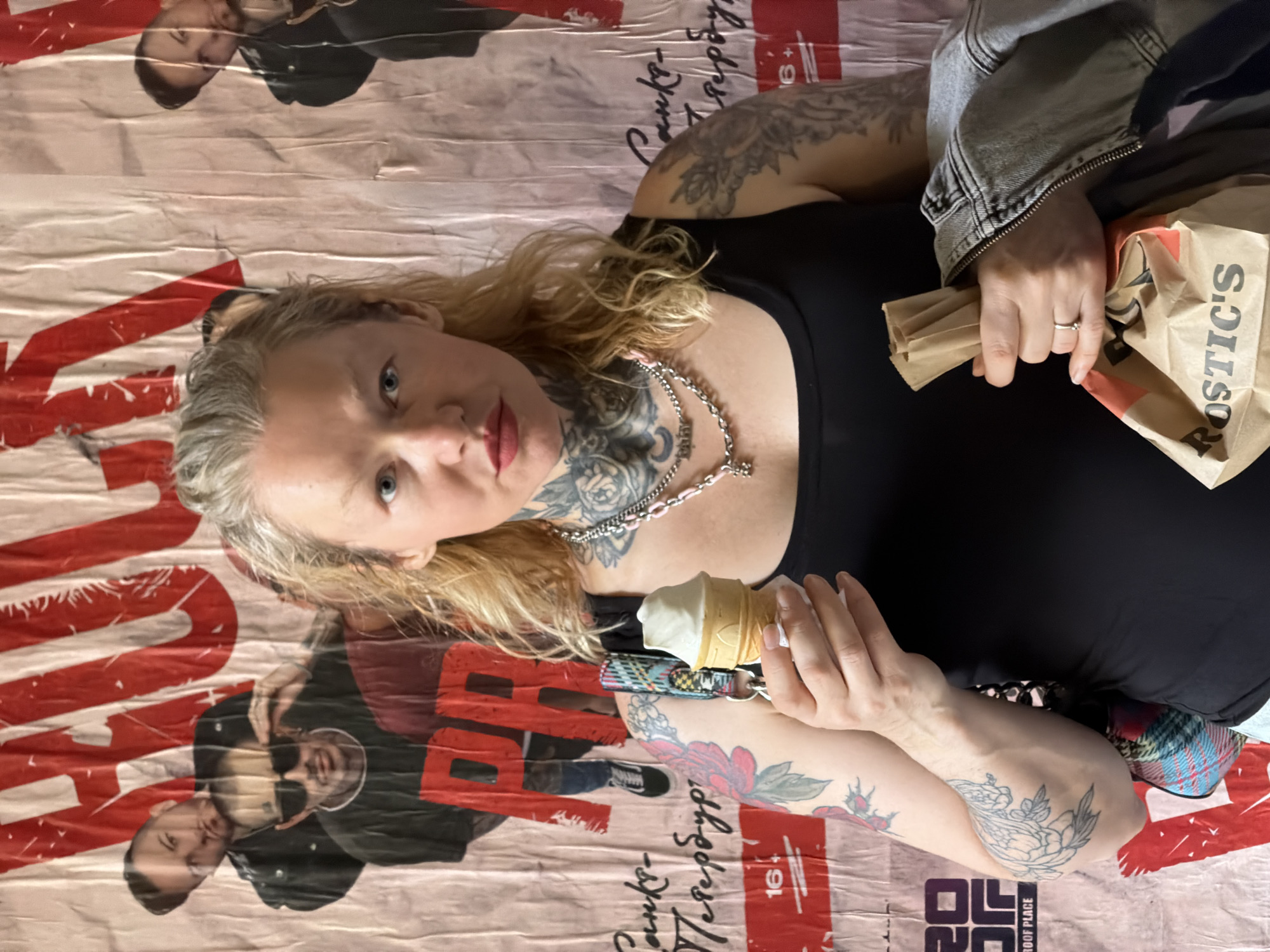 A blonde woman with extensive tattoos on her neck, arms, and chest stands in front of a large, crumpled poster. The poster appears to be a promotional or artistic piece with red and white elements and images of men. The woman is wearing a black tank top and a denim jacket draped over her shoulder. She is holding a small ice cream cone in her left hand and a brown paper bag in her right hand. The bag has the word "ROSTIC'S" printed on it. The background poster has text that appears to be in Russian, and includes the number "16+". The overall scene suggests a candid moment possibly in an urban environment. The presence of the Rosti's bag implies the location could be a place where this fast food chain is available.