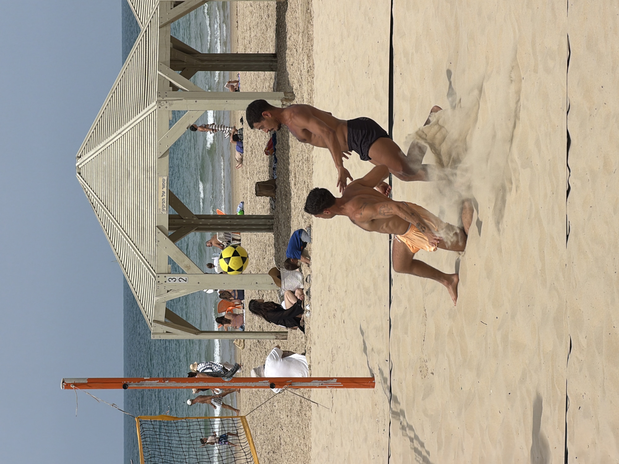 The image captures a beach volleyball game in Tel Aviv, Israel. Two shirtless men are actively engaged in the game, with one diving towards the sand and kicking up dust. In the background, there's a beach hut with people resting underneath. A volleyball net is visible in the foreground, along with beachgoers in the distance. The sky is clear.