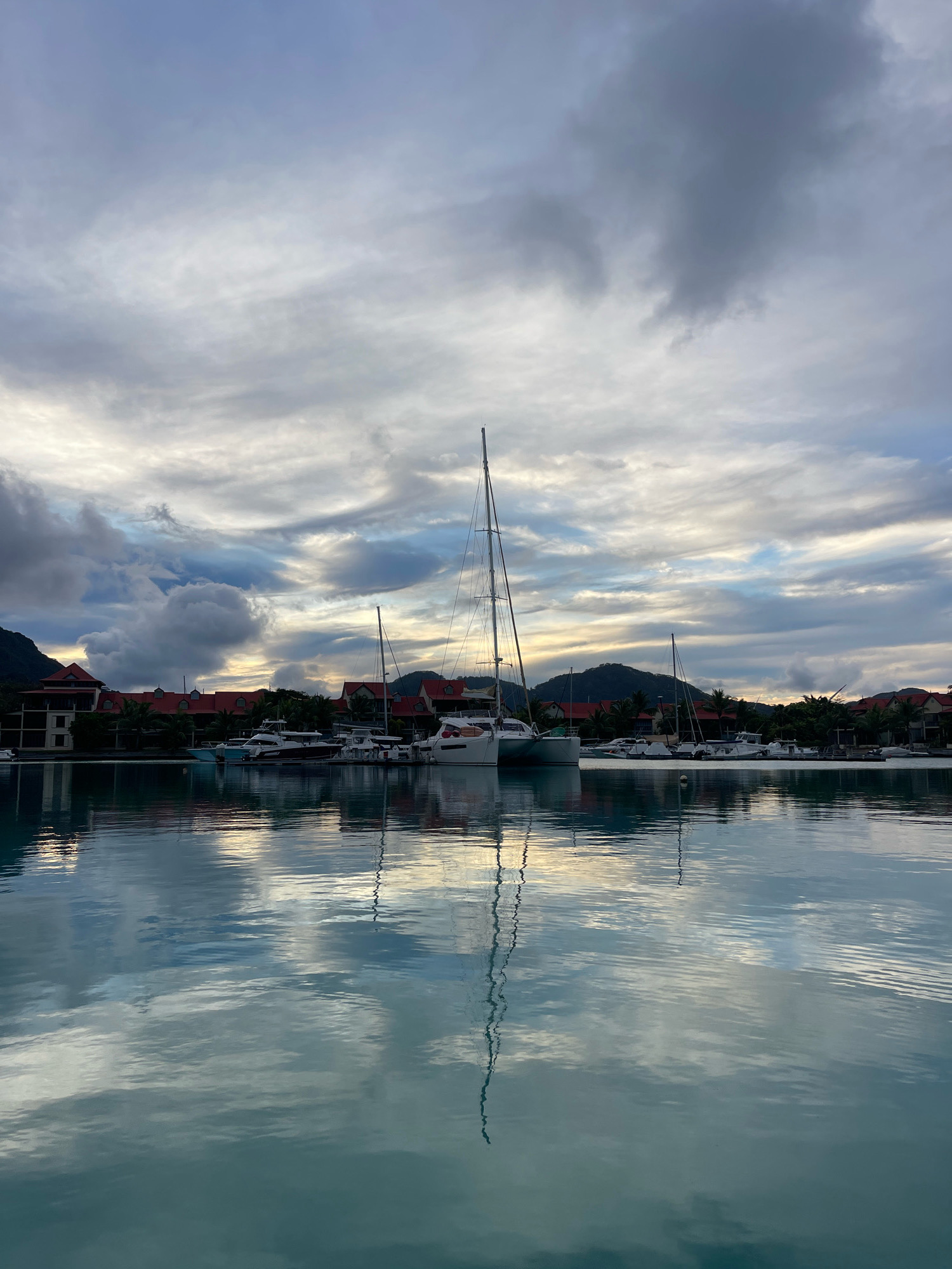 The image depicts a serene harbor scene in Providence, Seychelles. The foreground is dominated by a calm, reflective body of water, showing a mirror-like surface that captures the sky and the moored boats. In the midground, several sailboats and motor yachts are docked along a pier. The boats vary in size and design, with prominent masts and superstructures visible. Behind the boats, a strip of lush green vegetation and tropical trees lines the waterfront. Further back, a complex of buildings with distinctive red-tiled roofs is nestled against a backdrop of undulating hills. The sky above is filled with a dramatic display of clouds, ranging from light, wispy formations to darker, more substantial masses. The overall lighting suggests either dawn or dusk, with soft, diffused light casting a gentle glow over the scene. No people are visible in the image, and the atmosphere is one of tranquility and natural beauty.