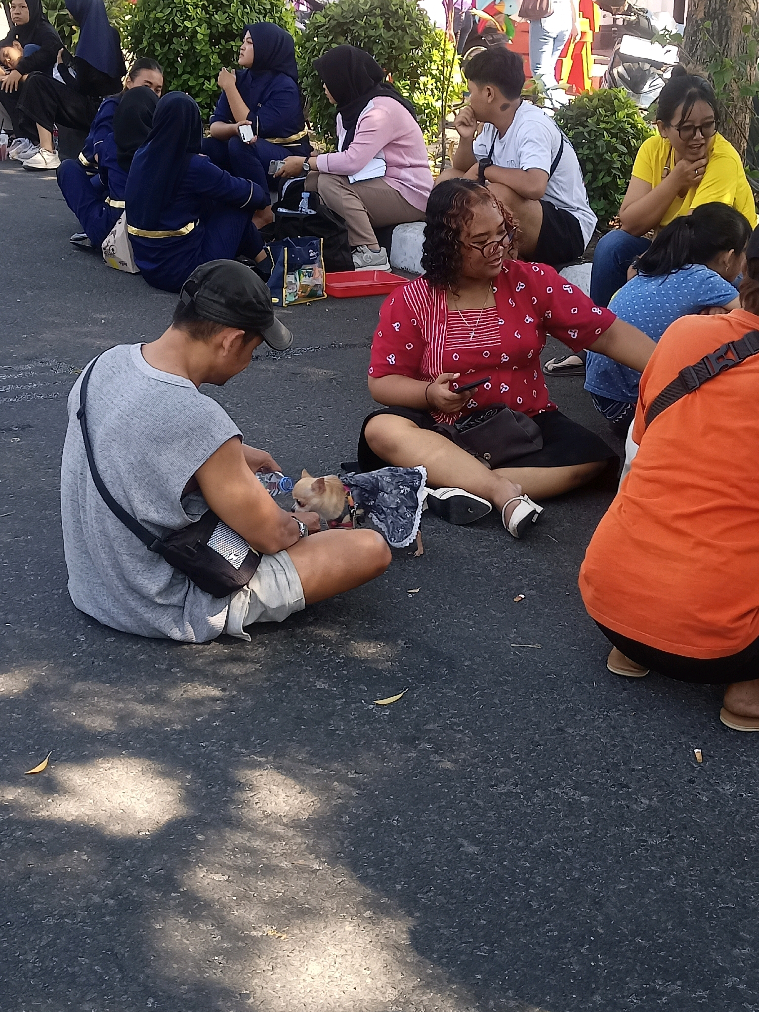In a sun-dappled outdoor setting, likely on asphalt, a group of people are seated on the ground. In the foreground, a person wearing a grey t-shirt and shorts is holding a small dog, offering it water from a bottle. The dog appears to be wearing a patterned vest. To their right, a person in a red and white striped top sits with a black bag and a mobile phone. Further back, several individuals in blue uniforms with gold trim are also seated, some interacting with each other. A person in a yellow shirt and another in a pink shirt are visible among them. The background features greenery, suggesting a park or outdoor event area, possibly near the vicinity of the Stadion Sriwedari in Surakarta, Indonesia, given the cultural attire and common gathering spots in the city. The overall atmosphere appears relaxed, with people waiting or taking a break.