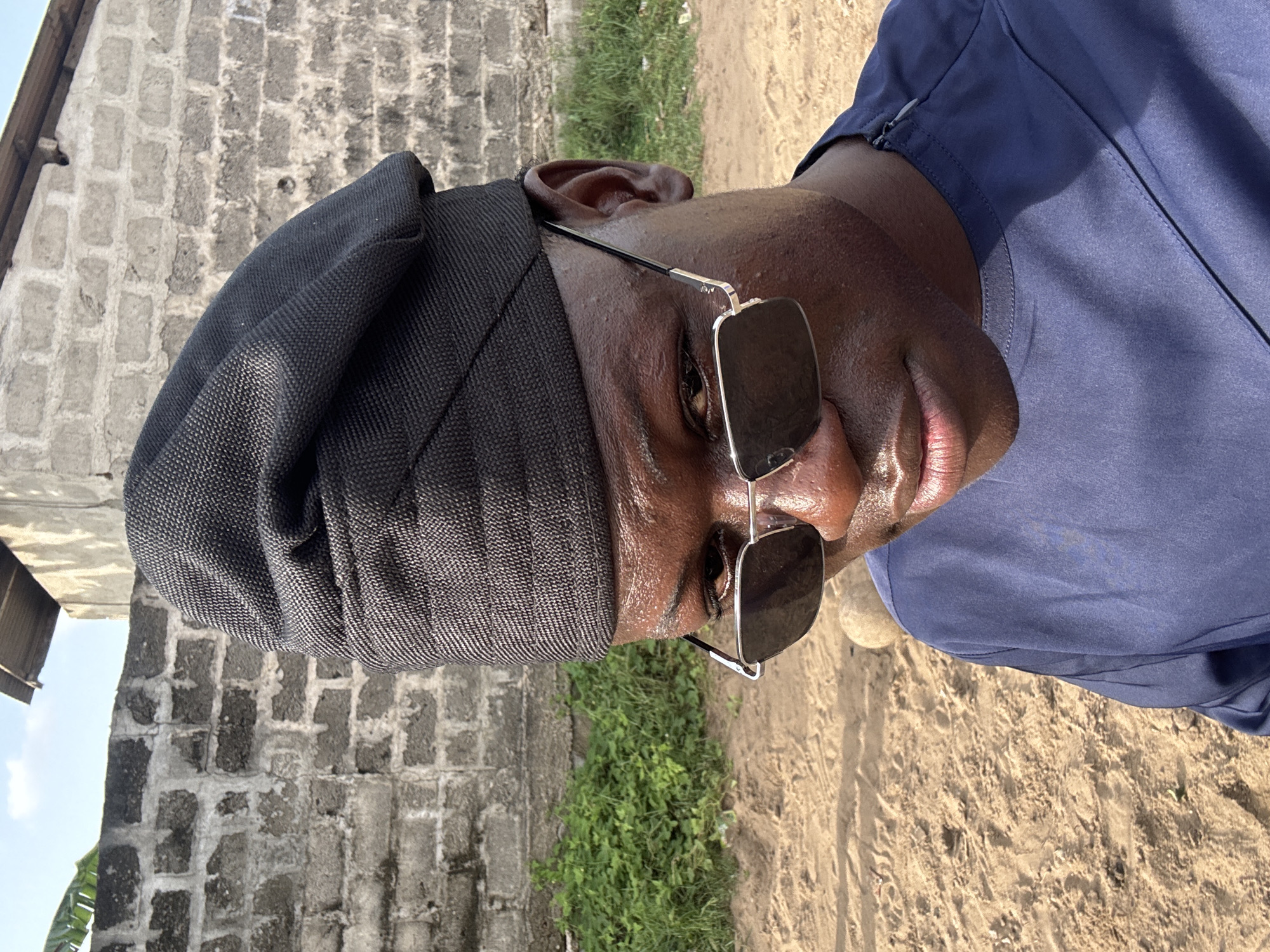 A man is pictured in Badagry, Nigeria. He wears a black cap and sunglasses with silver frames. He is wearing a blue shirt. He is standing on sandy ground with some green foliage. Behind him is a grey block wall.