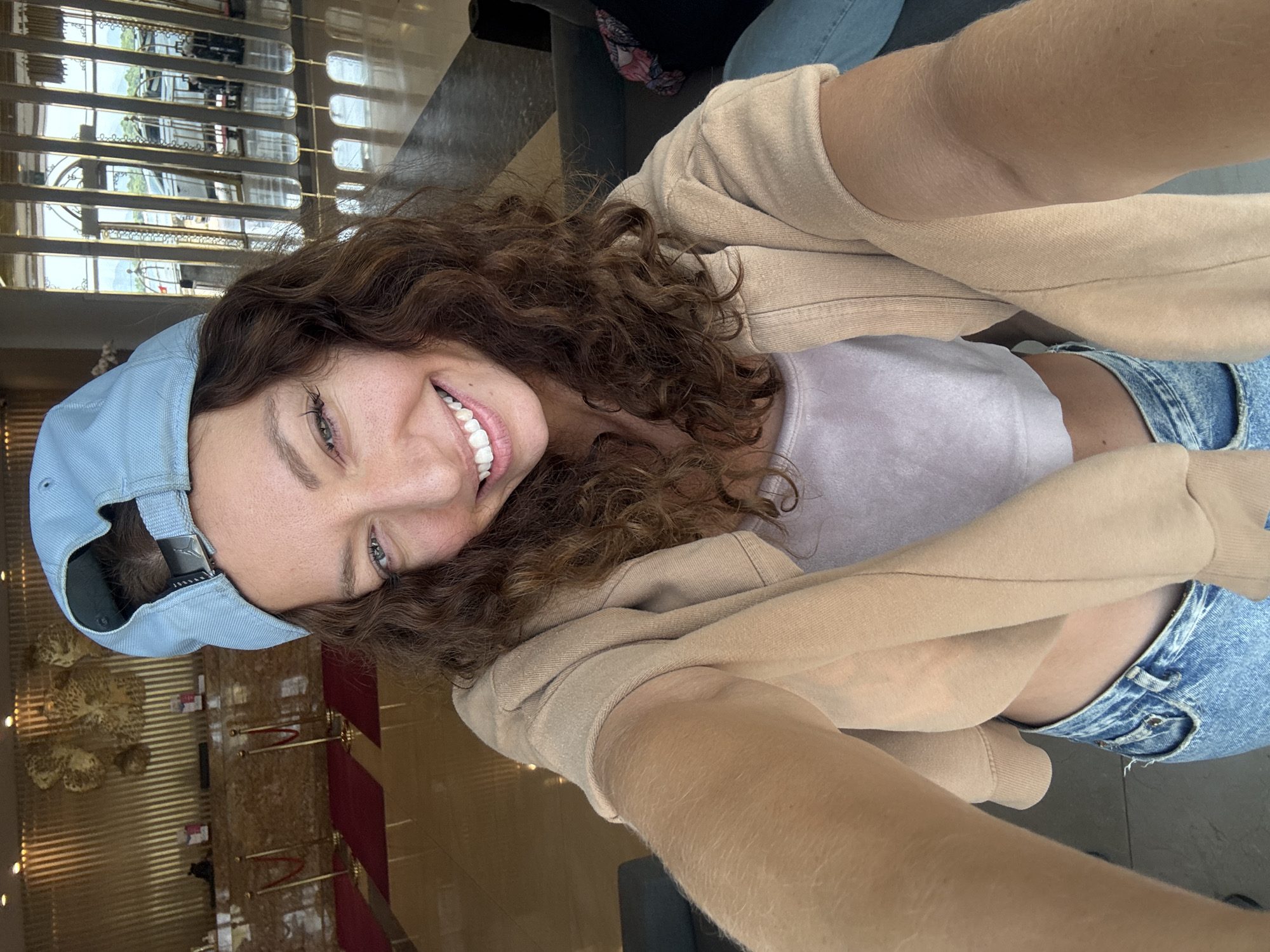 A woman is taking a selfie indoors. She is wearing a light blue baseball cap, a light purple top, a beige zip-up hoodie, and light blue jeans. She has curly brown hair and is smiling. The background appears to be a hotel lobby in Đà Nẵng, Vietnam. Features include decorative elements on the wall, metal railings, and reflective surfaces.