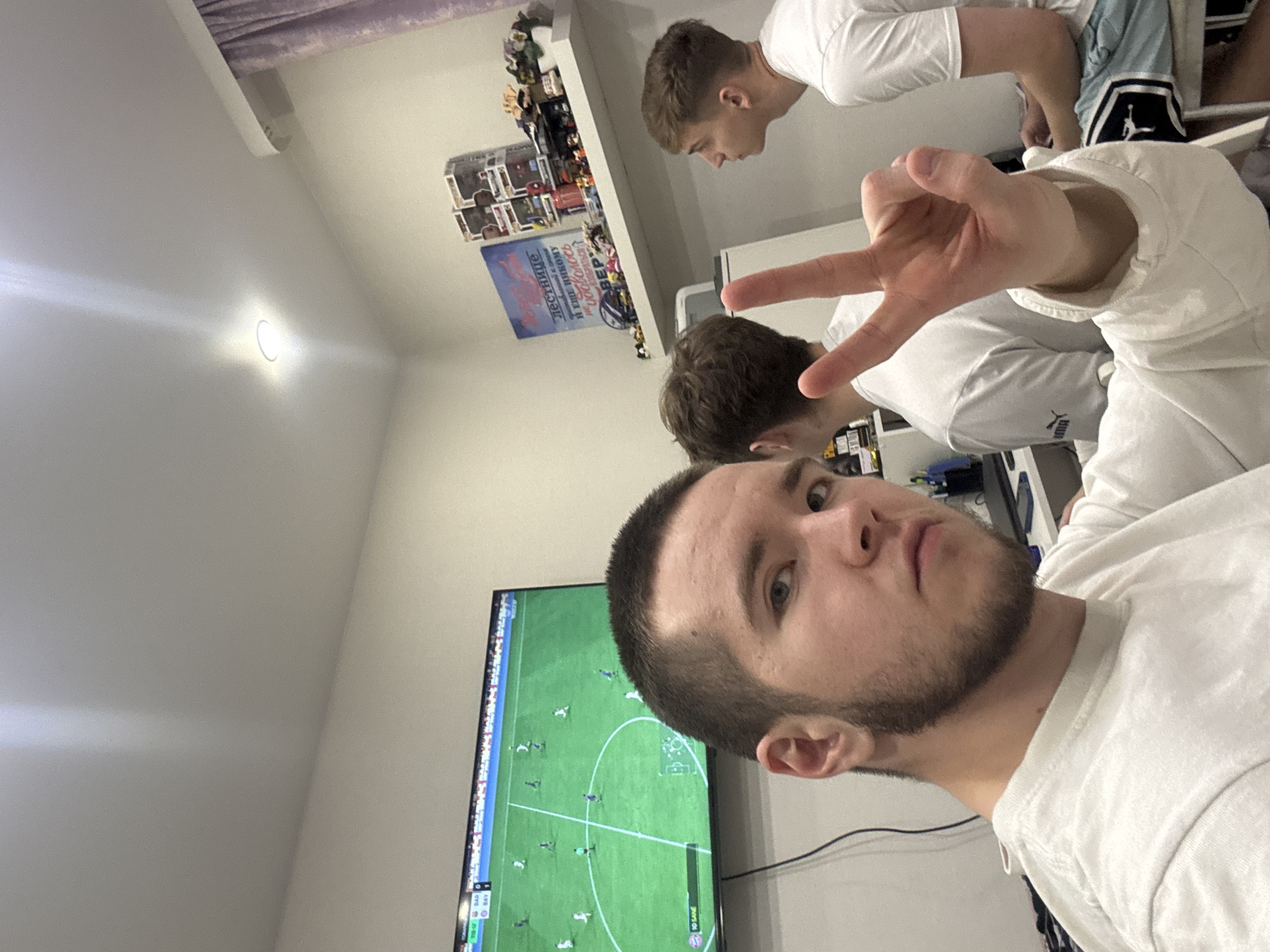 Three young men are captured in a casual indoor setting, likely in Kazan, Russia, as one of them takes a selfie. The man in the foreground, with short dark hair and a beard, looks directly at the camera with a neutral expression, holding up a peace sign with his right hand. He is wearing a white long-sleeved top. Behind him, another man in a white t-shirt, possibly bearing a Puma logo, is partially visible, appearing to be engaged with the TV screen. Further in the background, a third man in a white t-shirt and light-colored shorts with a Jumpman logo (Jordan brand) leans over a surface.
The central activity appears to be watching or playing a soccer video game on a large flat-screen television, which displays a match between "SAD" and "BAY" with a "0-0" score at "18:07" game time. The room has white walls and is lit by an overhead circular light fixture, suggesting it might be evening or a dimly lit interior. A shelf on the back wall holds numerous collectibles, possibly Funko Pop figures, and below it, a poster with Russian text is visible, including "ДЕСЯТИЛЕТИЕ" (Decade) and "РЕЗЕРВ" (Reserve). The overall atmosphere is relaxed and informal.