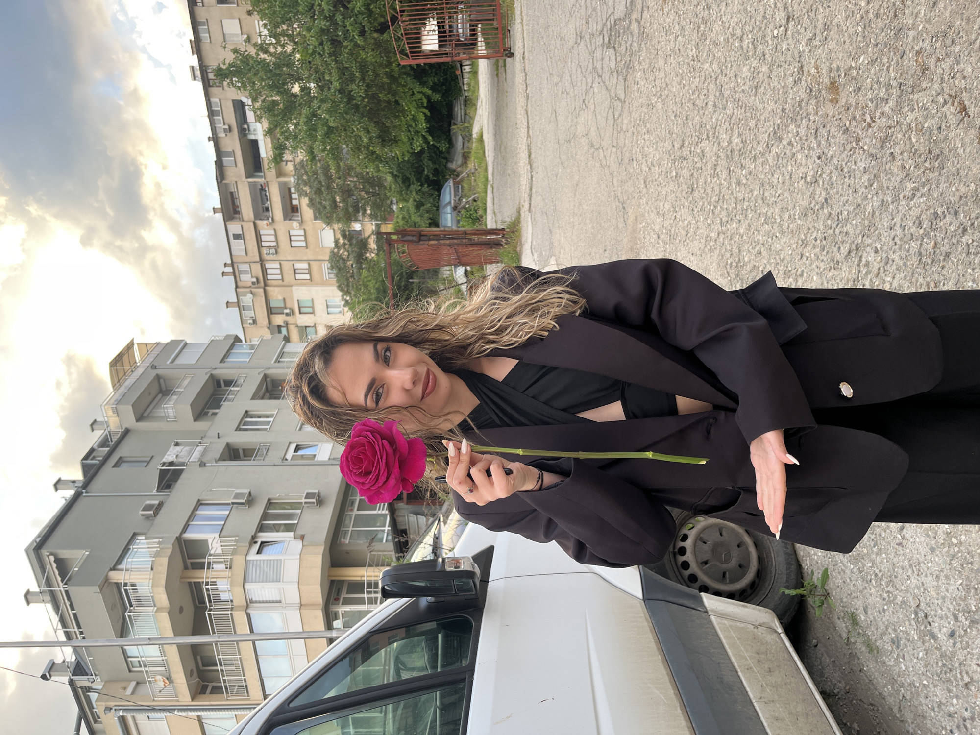 A young woman with blonde curly hair is holding a pink rose in her left hand, standing next to a white van on a street in Blagoevgrad, Bulgaria. She is wearing a black blazer and a black top. Behind her, there are several residential buildings, trees, and a metal gate. The street is paved with cracked asphalt. A car tire is visible near the van.