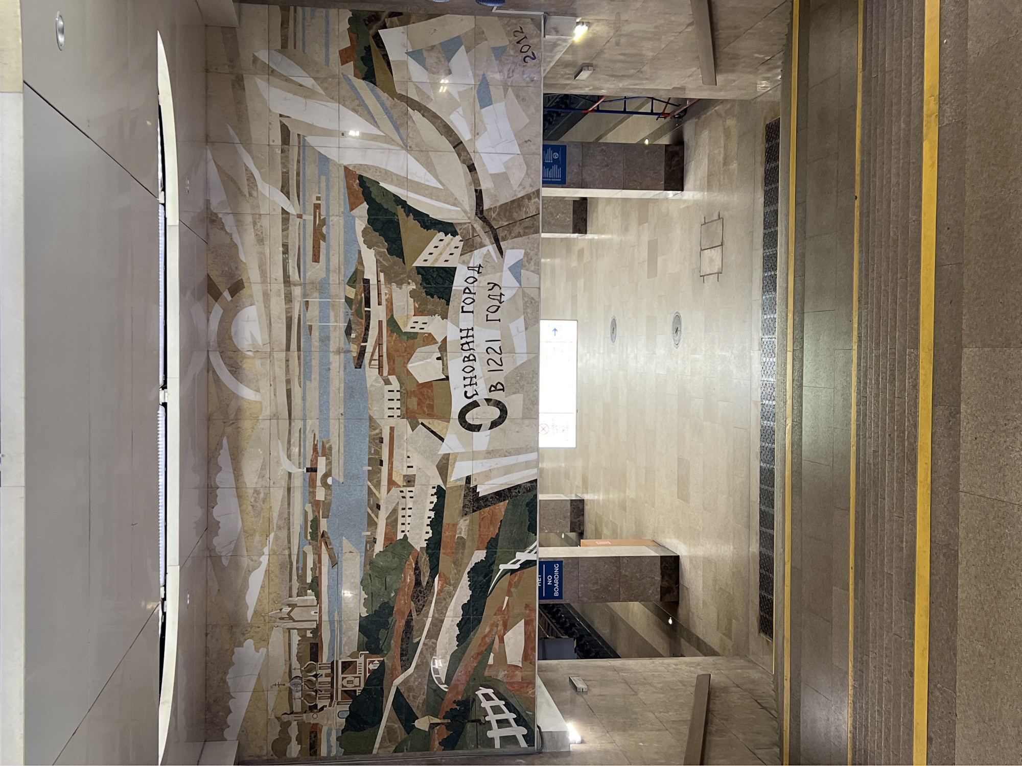 The image shows a wide mosaic mural decorating a wall in what appears to be a public transit station, likely a metro station, in Nizhny Novgorod, Russia. The mural depicts a city landscape with domed buildings, a river, and stylized natural elements. Text in Russian is integrated into the mural, reading "ОСНОВАН ГОРОД В 1221 ГОДУ" (The city was founded in 1221) and "2012". Below the mural, a walkway leads away, with stairs visible in the foreground and on the sides. The architecture is characterized by beige marble or stone finishes. Yellow safety lines mark the edge of the stairs. A "NO BOARDING" sign is visible on a pillar to the left. The environment is clean and well-lit, typical of a modern underground transportation hub. There are no people clearly visible in the frame.