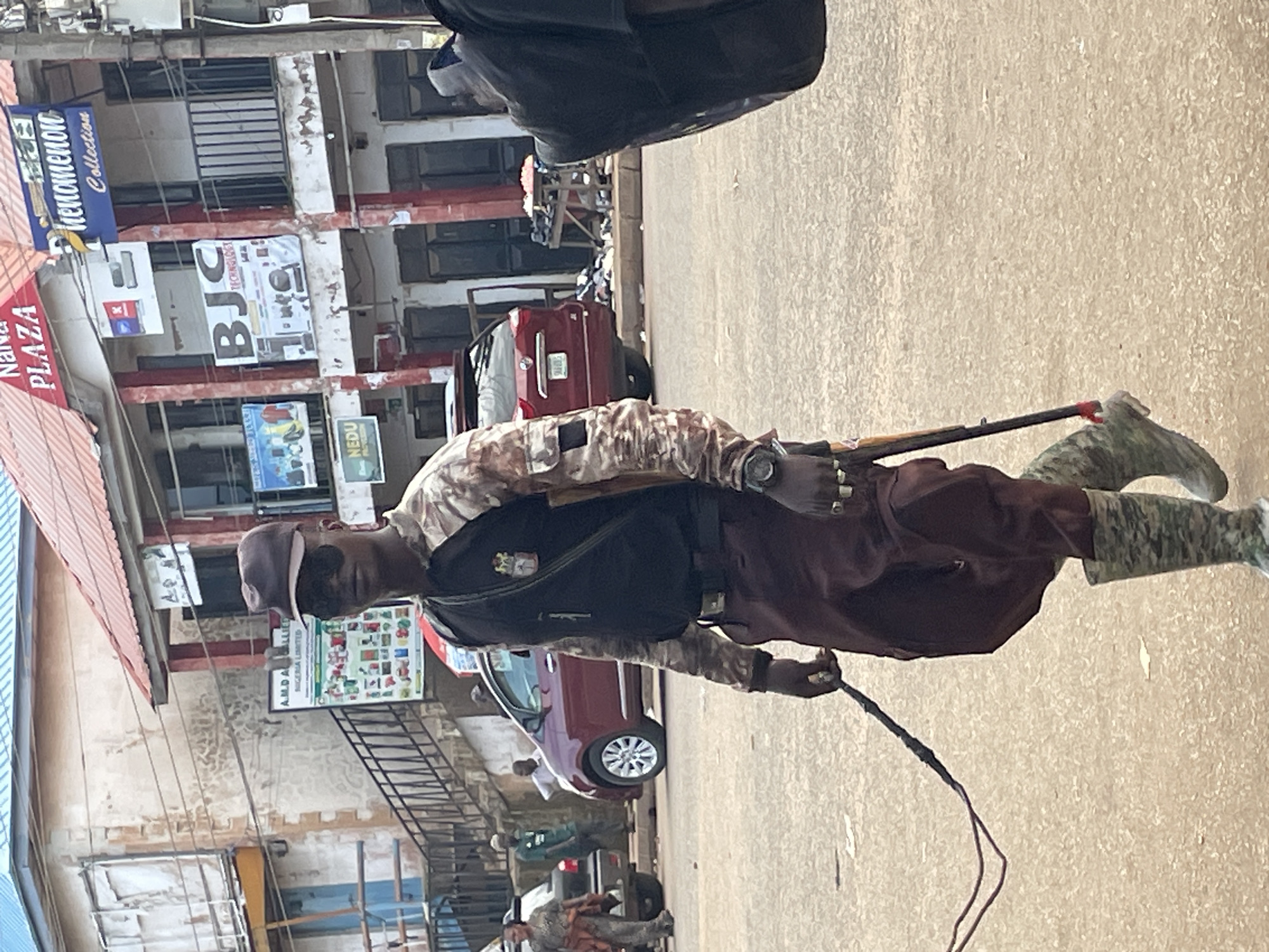A person dressed in camouflage military attire, including a cap, vest, and patterned boots, stands in the street holding what appears to be a rifle. The person also holds a whip in their right hand. The scene is set on a street in Bukuru, Nigeria, with buildings lining the road. Signs on the buildings include "NANA PLAZA," "BJC TECHNOLOGY," and "A.M.D.A ALLIES NIGERIA LIMITED." A red car and other vehicles are visible in the background, along with some market stalls. The lighting suggests it is daytime, and the weather appears to be fair. The person's posture and the items they are holding suggest a sense of authority or control, though no specific event is clearly depicted. The overall impression is a street scene in a Nigerian town with a person in military-like attire present.