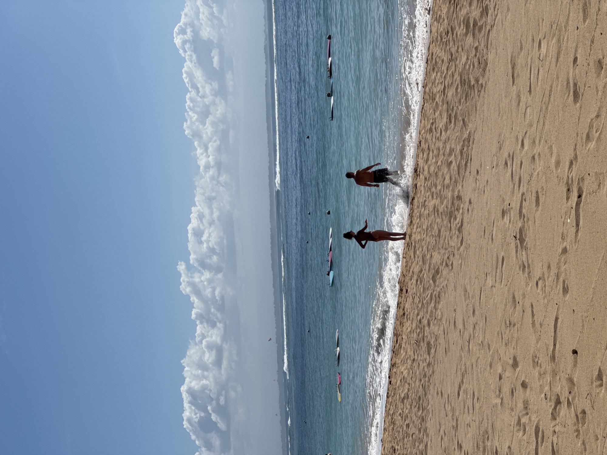 The image depicts a beach scene in Kuta Selatan, Indonesia. The sky is predominantly blue with large white cumulus clouds. The ocean transitions from a lighter turquoise near the shore to a deeper blue further out. Several individuals and surfboards are visible in the water. Two people, silhouetted against the light, stand on the beach near the water's edge. One appears to be male and is partially submerged in the shallow waves. The other appears to be female and is standing on the sand. The beach itself is a light tan color, marked with numerous footprints. The perspective is from a slightly elevated position on the sand, looking out over the water.