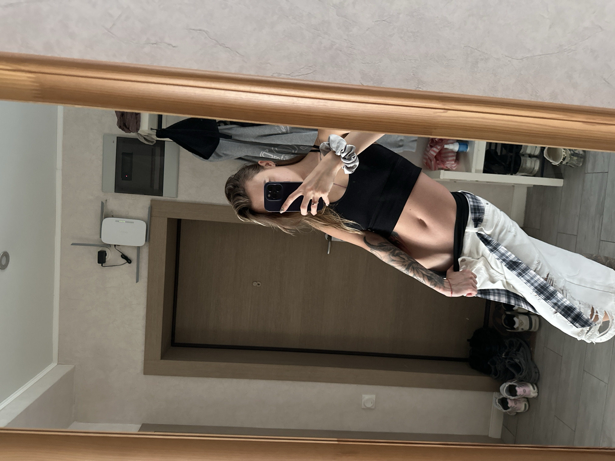 A young Caucasian woman with blonde hair tied in a ponytail is taking a mirror selfie in what appears to be an apartment hallway. She is wearing a black crop top and white ripped jeans with a plaid accent. Her left arm is covered in tattoos. In the reflection, a wooden door with a silver handle is visible, as well as a shelf with shoes and a bottle of spray on the right. The floor is tiled and there are several pairs of sneakers on the floor. On the wall, a white router is mounted. The lighting suggests it is daytime.