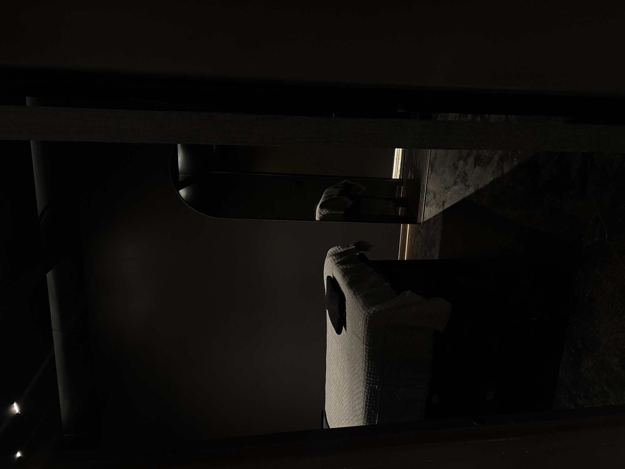 The image shows a dimly lit interior space in Ciudad López Mateos, Mexico. There is a cylindrical object to the left, possibly a pipe or post. Above, there is a reflective surface with an indistinct object on it. A box-shaped object with a patterned surface is visible in the foreground to the right, and it appears to have a dark opening on one side. The environment is dark, and the specific details are hard to distinguish.