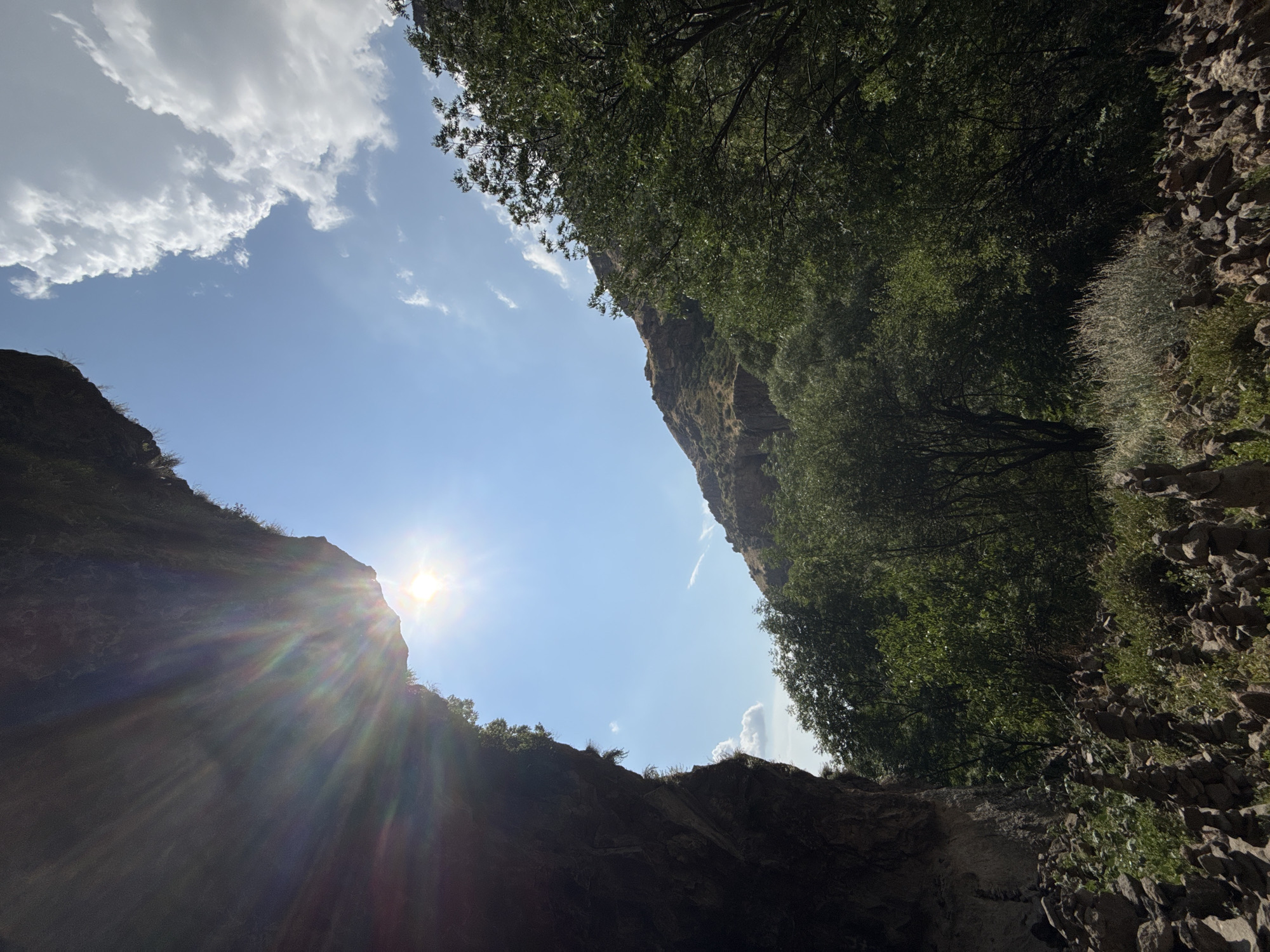 The image depicts a view from within a rock overhang or cave entrance, looking out towards a bright blue sky with scattered white clouds. The sun is prominently visible, creating a lens flare effect with visible light rays. On the left side of the frame, a dark, rocky overhang dominates the foreground, with some vegetation growing on its edge.
Beyond the overhang, a steep, rocky hillside covered in sparse vegetation rises. The middle ground is filled with dense green trees, suggesting a forested area. In the lower foreground, directly beneath the overhang, there are numerous small cairns or stacks of stones. These are arranged in clusters and appear to be man-made.
The scene is set in the natural environment of Goght, Armenia, specifically likely near the Geghard Monastery, which is known for its rock-cut architecture and the natural cave-like structures in the vicinity. The overall impression is one of a rugged, natural landscape bathed in sunlight. There are no people or animals visible in the image.