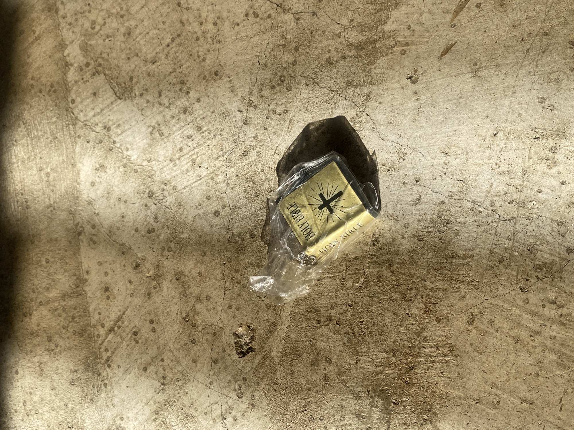 A small, gold-colored book, identified as a "Holy Bible," lies on a textured, light-brown concrete or paved surface. The book is encased in clear plastic, which appears somewhat crinkled and torn, particularly around its edges.
The cover of the book features the words "HOLY BIBLE" printed vertically along its visible spine, and a prominent black cross design with radiating lines on its face. The text and design appear slightly distressed.
The setting is an outdoor area, likely during the daytime, as strong sunlight casts a distinct, sharp shadow from the book and its plastic wrapping onto the ground. The ground itself is characterized by various cracks, small dark speckles, and an uneven, rough texture. No other objects or people are visible in the frame. The overall impression is of an item placed or discarded on a weathered surface. While the exact location cues are limited to the generic nature of the concrete, the conditions are consistent with an outdoor environment in a place like Davao City, Philippines.