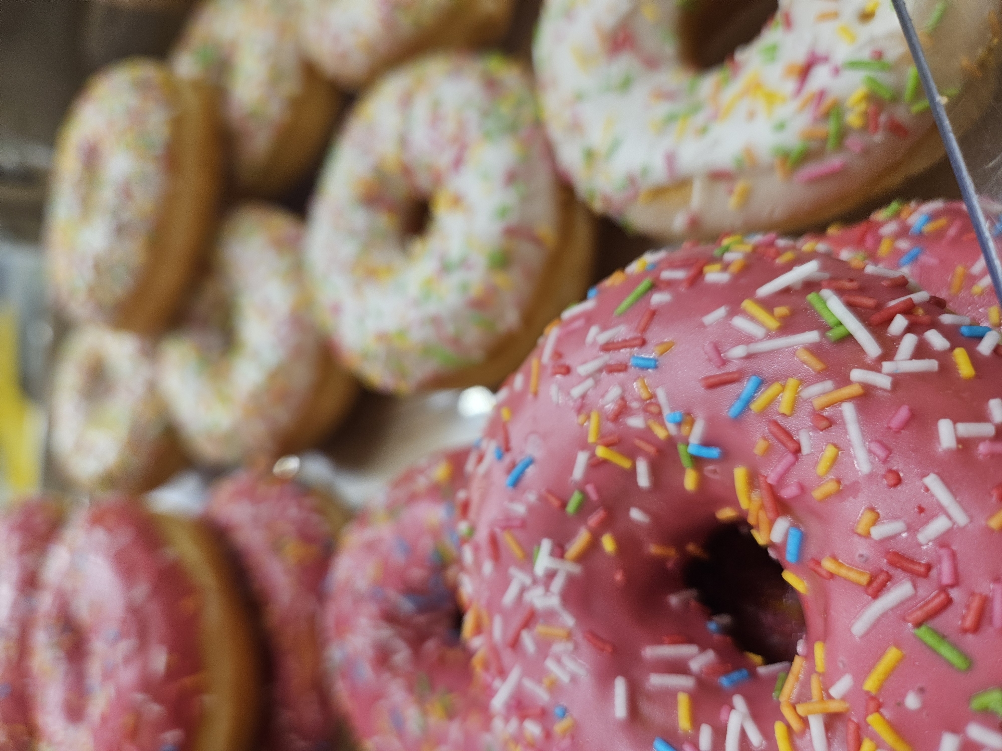 The image shows a close-up view of several donuts displayed in a clear plastic container, likely at a bakery or supermarket in Rostov-on-Don, Russia. The donuts are ring-shaped and topped with frosting and colorful sprinkles. There are at least two prominent pink-frosted donuts with white, yellow, blue, orange, and green sprinkles, and several white-frosted donuts with similar sprinkles. The donuts appear to be stacked or arranged in a way that suggests they are for sale. The lighting is bright, highlighting the textures and colors of the pastries. The background is blurred, but hints at a retail environment. There are no people visible in the image.
