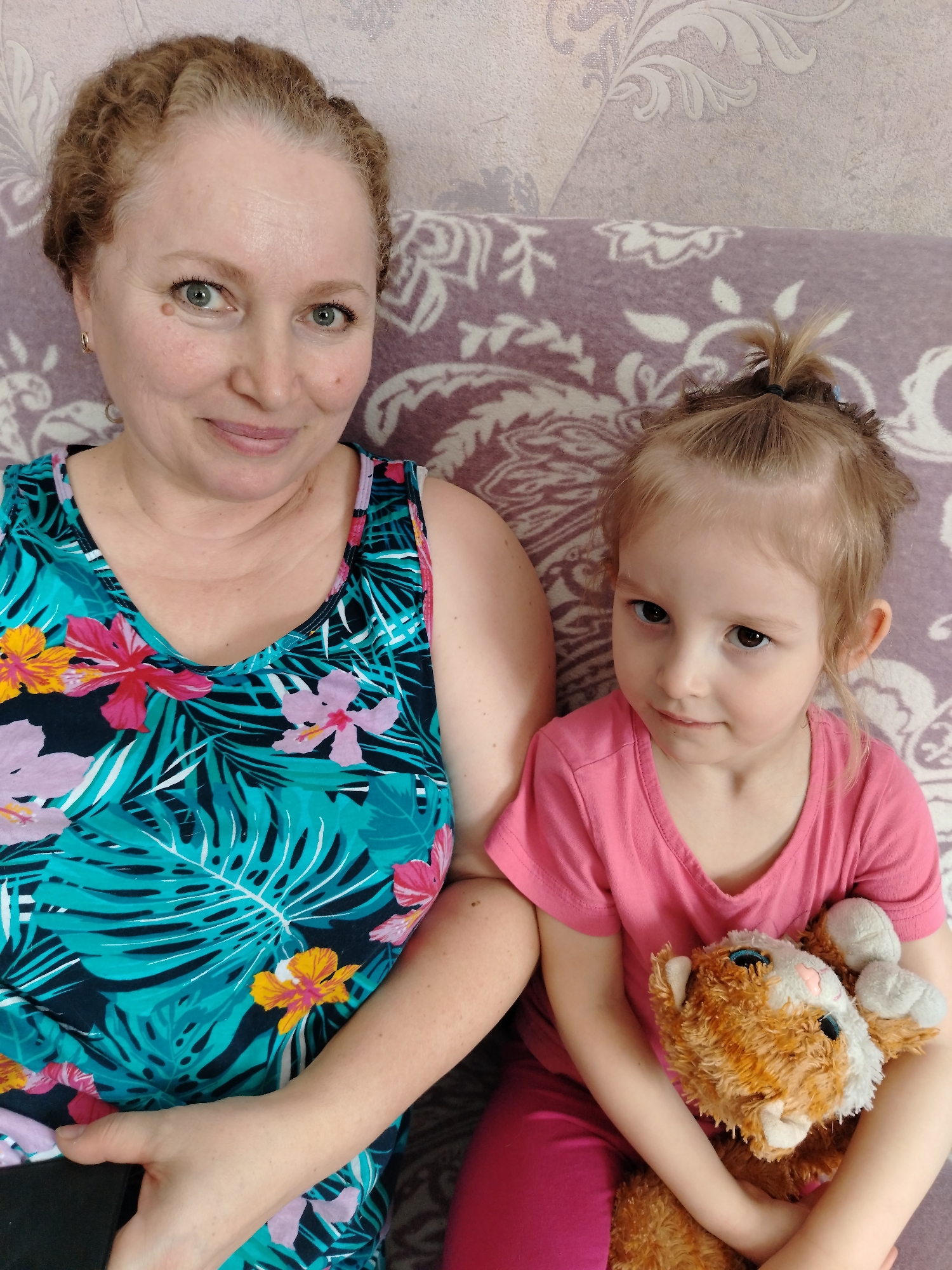 An adult female and a young female child are seated side-by-side in an indoor setting within Nizhnyaya Gar', Russia. They are positioned against a wall covered with light-toned, patterned wallpaper and are seated on a surface draped with a light purple patterned blanket or throw.
The adult female, with light reddish-brown hair styled in braids or curls, green eyes, and fair skin, is on the left. She wears a sleeveless top featuring a teal background with a vibrant pattern of pink, purple, and yellow flowers, and dark green/black tropical leaves. She looks directly at the camera with a slight smile. Her left arm is visible, and she holds a dark rectangular object, possibly a mobile phone.
The young child, appearing to be a girl aged approximately 3-5 years old, is seated on the right. She has light brown hair tied into a small ponytail at the crown of her head and dark brown eyes. She wears a plain pink short-sleeved t-shirt with a V-neck and pink pants. She looks slightly to her left (viewer's right) with a neutral expression, holding a brown and white plush toy with light blue eyes in both arms against her body.