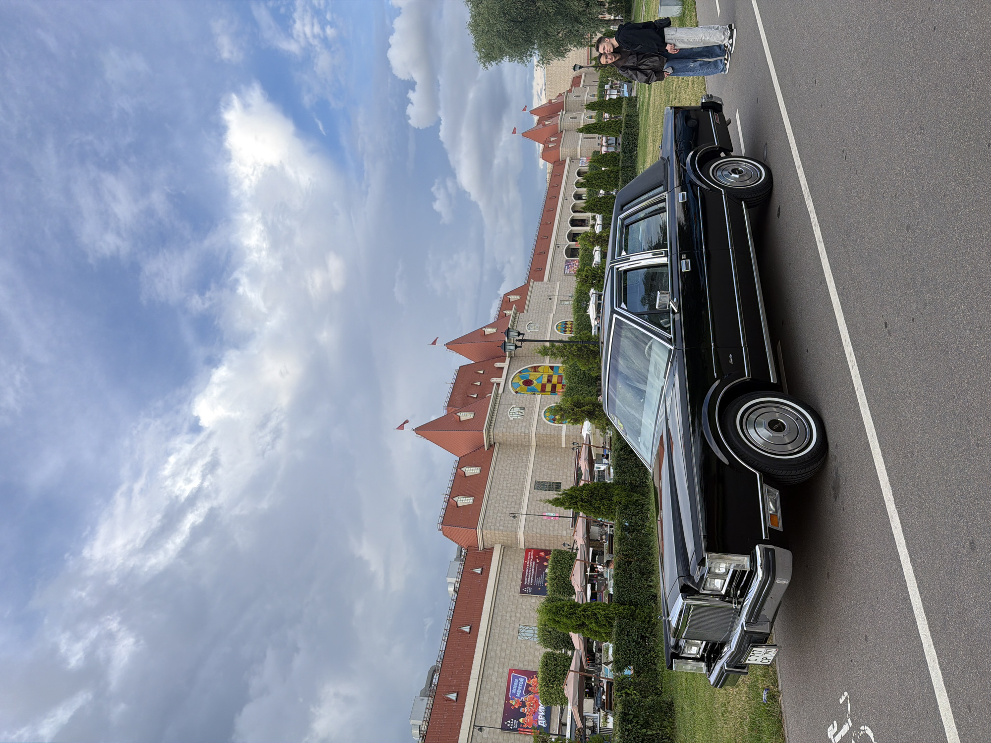 A black Lincoln Town Car is parked on an asphalt road with a white line. Two young people, a male and a female, stand to the right of the car, facing forward. The building in the background is a large, multi-story structure with red-tiled roofs and light-colored stone walls. There are tables and umbrellas set up in front of the building, suggesting an outdoor seating area. Trees and manicured bushes are visible in the foreground and along the building's facade. The sky is partly cloudy with a mix of gray and blue hues.