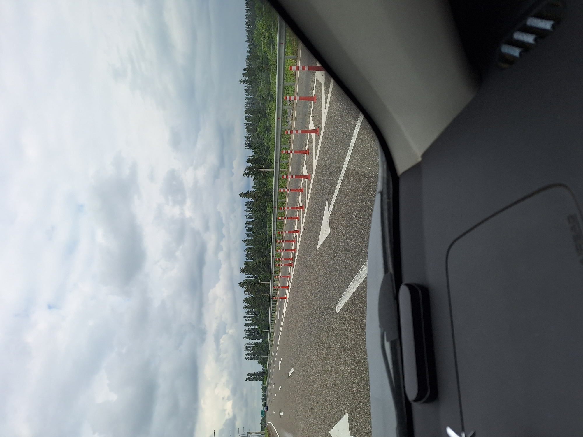 The image shows a view from the inside of a car driving on a highway. The highway has multiple lanes marked with white arrows, and on the right side, there is a row of orange traffic cones with white reflective stripes. Beyond the highway, there is a metal guardrail, followed by a line of tall, slender trees forming a dense forest. The sky is overcast with heavy clouds. In the foreground, part of the car's dashboard is visible, including a black oblong object on the dashboard and the edge of the windshield. A sign is visible in the distance on the left side of the highway. The location is identified as being in the city of Lanchkhuti, Georgia.