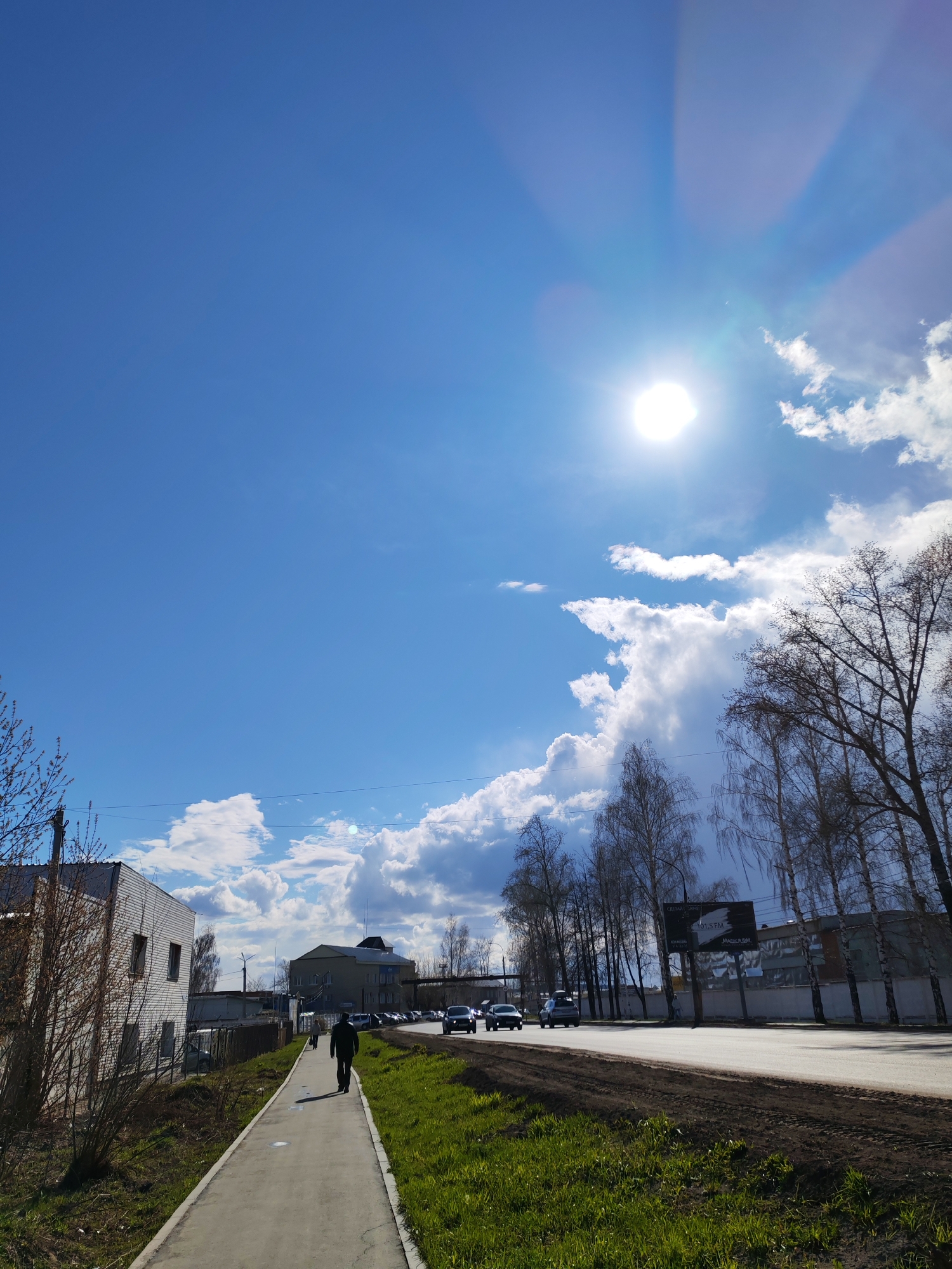 The image captures a sunny daytime scene on a street in Chaikovsky, Russia, likely during late winter or early spring, given the bare trees and emerging green grass. The bright sun is prominently featured in the upper right quadrant of the sky, casting lens flares across the blue expanse, which is dotted with scattered white clouds.
In the foreground, a paved sidewalk runs along the left side of the frame, bordered by grassy patches and sparse, small trees and bushes. A lone person, seen from behind, walks away from the camera on the sidewalk. To their left, there's a two-story white building with multiple windows.
Further into the background, a road stretches diagonally across the frame, with several cars visible, indicating ongoing traffic. A line of bare-branched trees, possibly birches, flanks the right side of the road and extends into the mid-ground. Among these trees, a large billboard is visible. It displays text in Cyrillic, including "ОТДЕЛЫ ЗАПЧАСТЕЙ" (Spare Parts Departments), "107.5 FM", and "МАРКС.РМ". In the distance, more buildings and vehicles can be seen, hinting at an urban or suburban environment. Power lines crisscross the upper part of the scene against the clear sky.