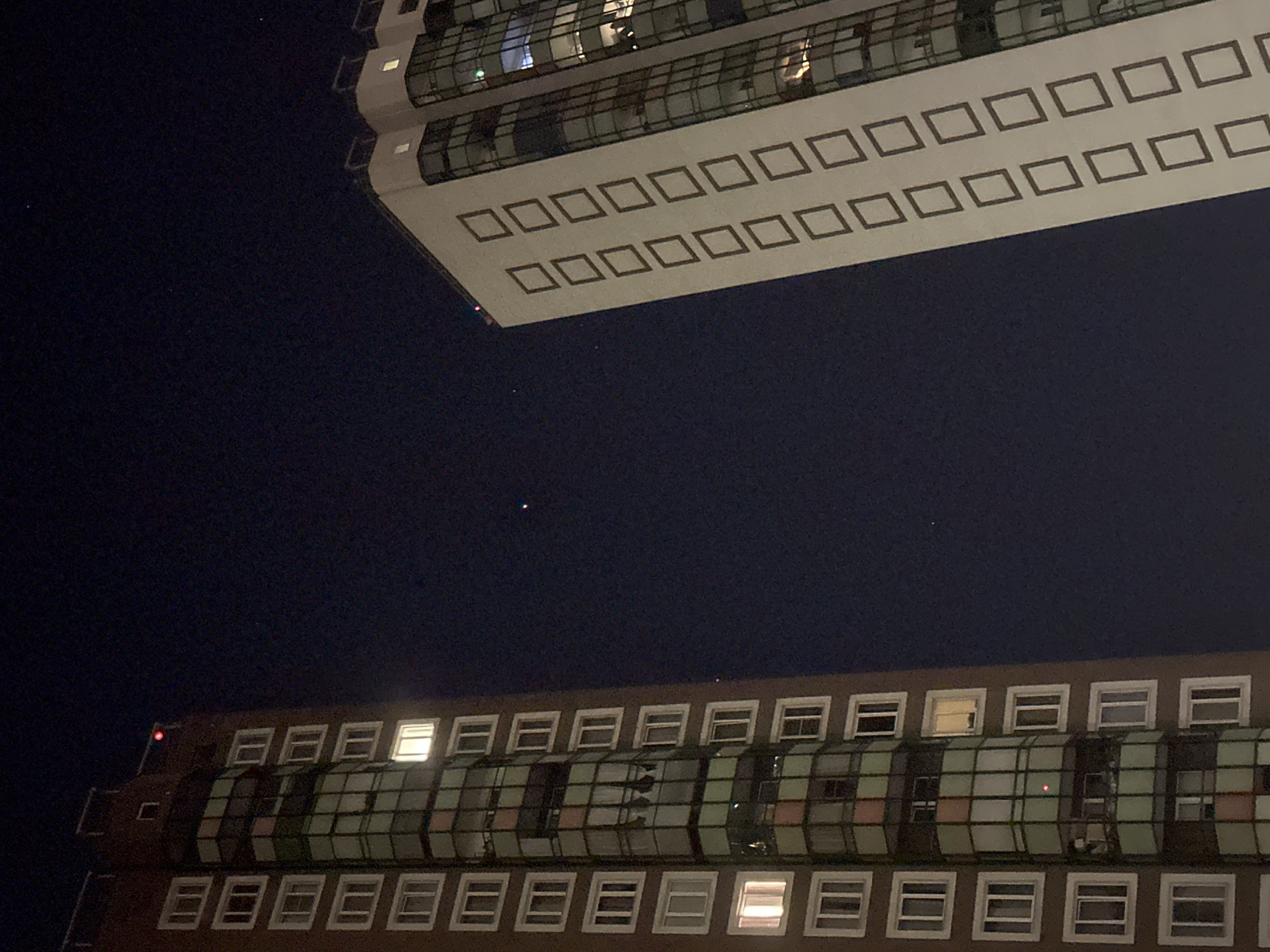 The image shows two tall buildings at night in Sinyavino, Russia. The building on the left is brown and has a series of rectangular windows, some of which are illuminated. It also has a curved section with colored panels. The building on the right is white and has a series of square decorative elements along its side. It has many glass windows that reflect the lights from inside. The sky is dark and appears to have a few faint stars or distant lights. There are no visible people in the image.