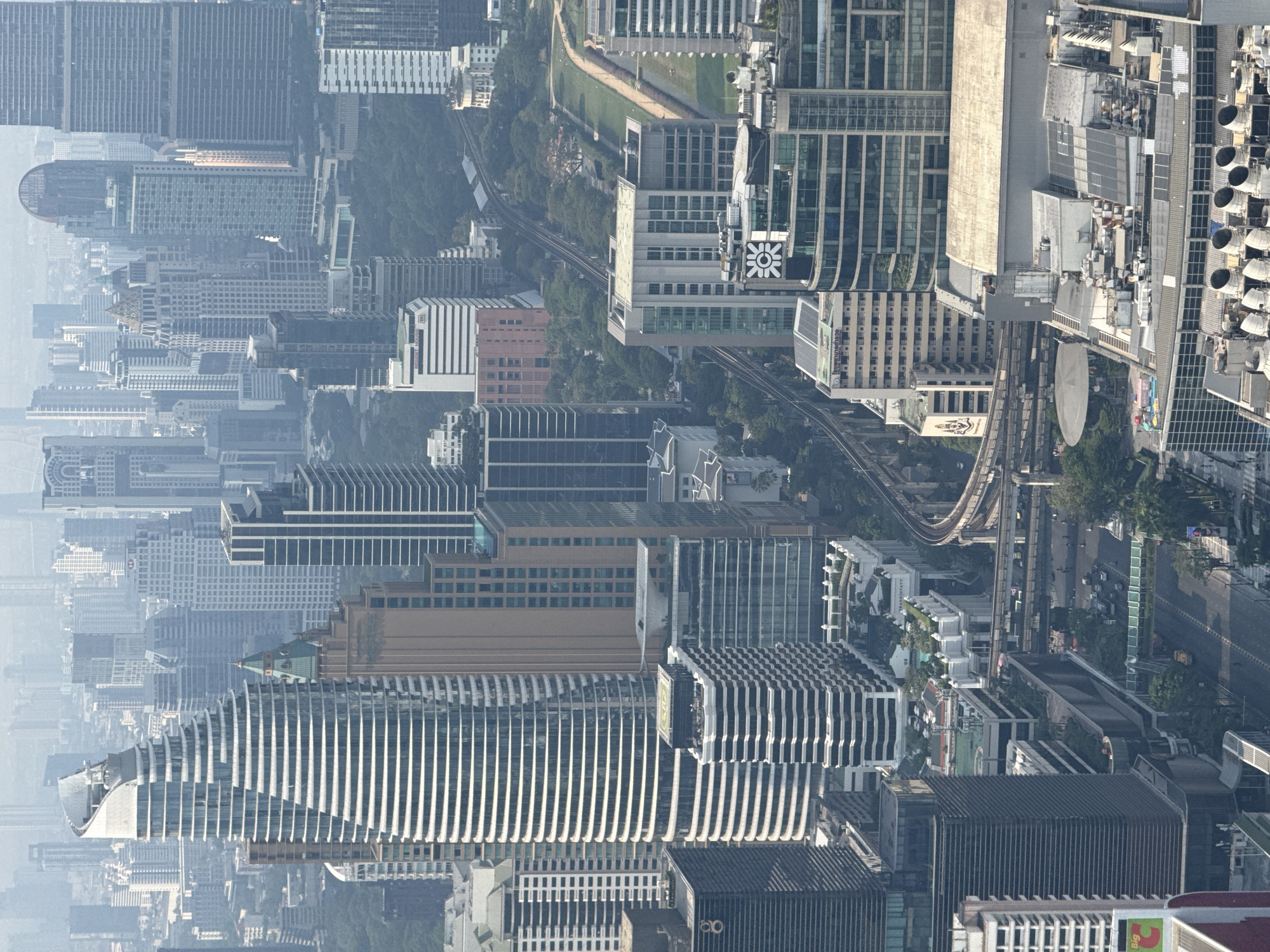 The image shows an aerial view of Bangkok, Thailand. It is taken from a high vantage point and shows a dense cityscape with numerous high-rise buildings. Many buildings have modern designs with vertical stripes. There is a haze over the city. A monorail track winds through the buildings. The image provides a glimpse into the urban environment, showcasing the city's architecture and infrastructure.