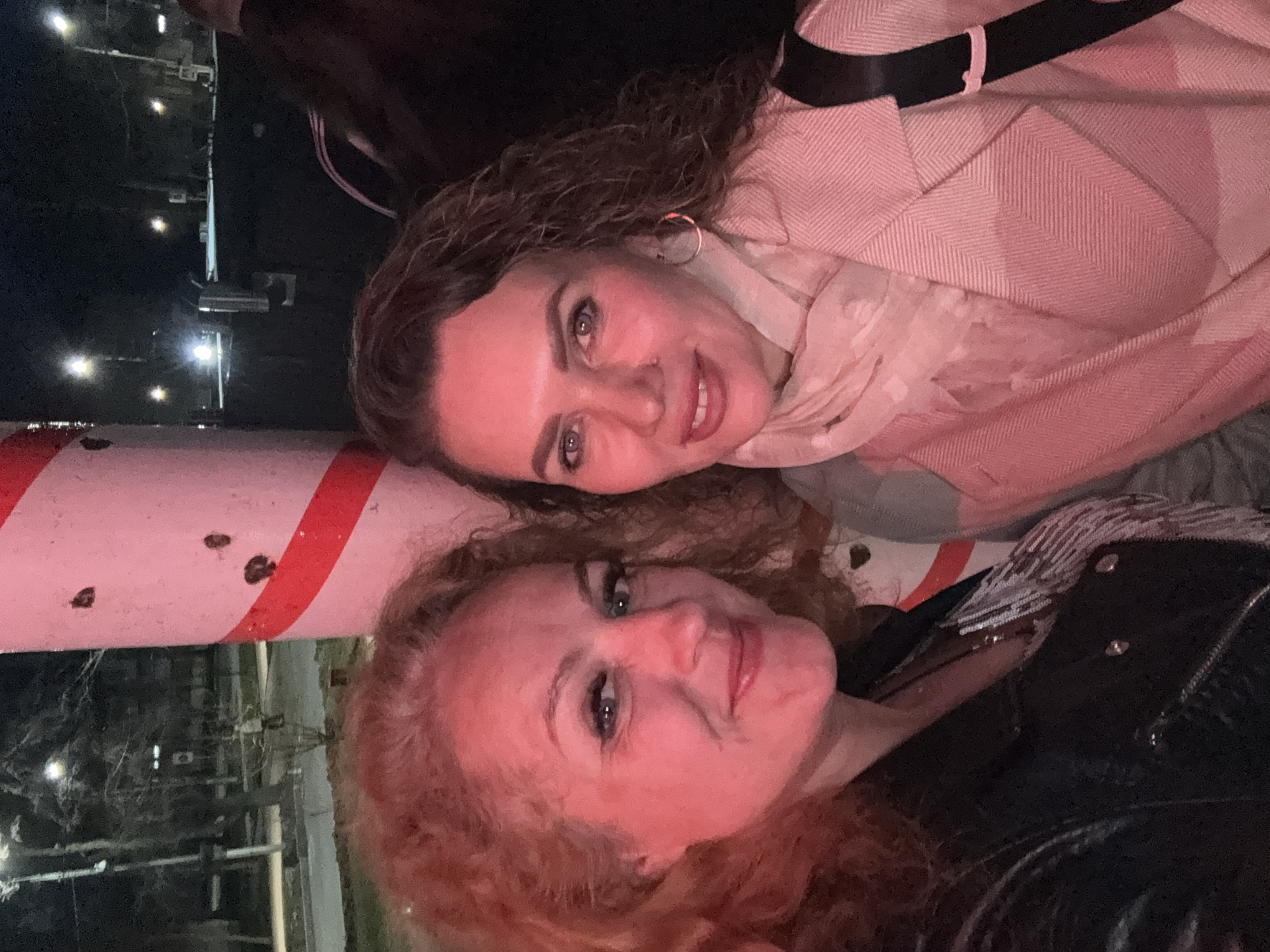 Two women pose cheek-to-cheek near a red and white striped pole at night. The woman at the top has brown, curly hair and wears a pink herringbone coat with a patterned scarf and silver hoop earrings. The other woman has reddish-blonde curly hair and wears a black leather jacket. Outside the city of Chaikovsky, Russia, lights and structures can be seen in the background.