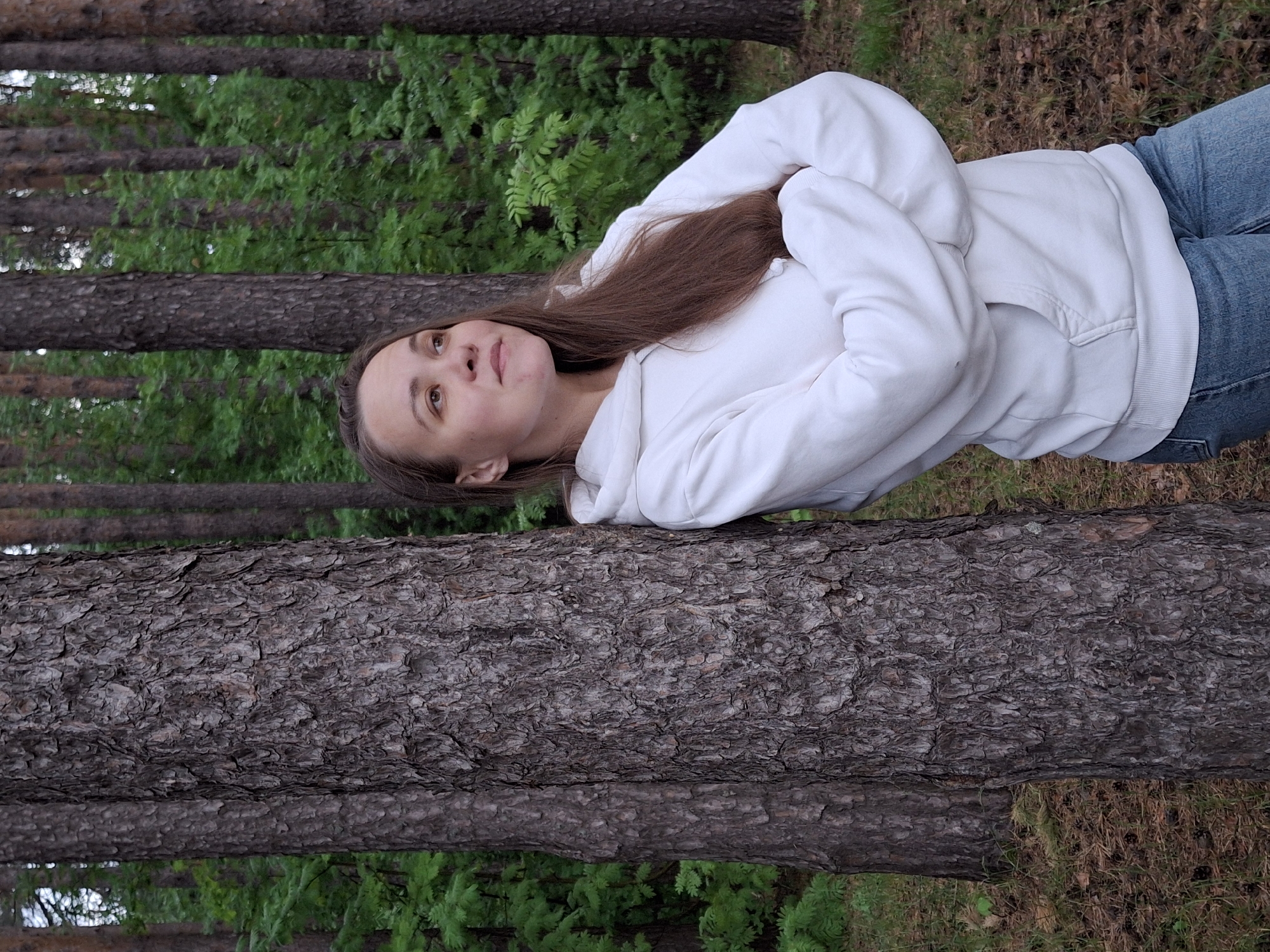 A fair-skinned woman with long brown hair lies on a tree trunk in a forest. She is wearing a white hooded sweatshirt and blue jeans. She has her arms crossed over her chest and is looking up. The forest has tall, thin trees with green ferns growing around their base. The location is implied as Chaikovsky, Russia.