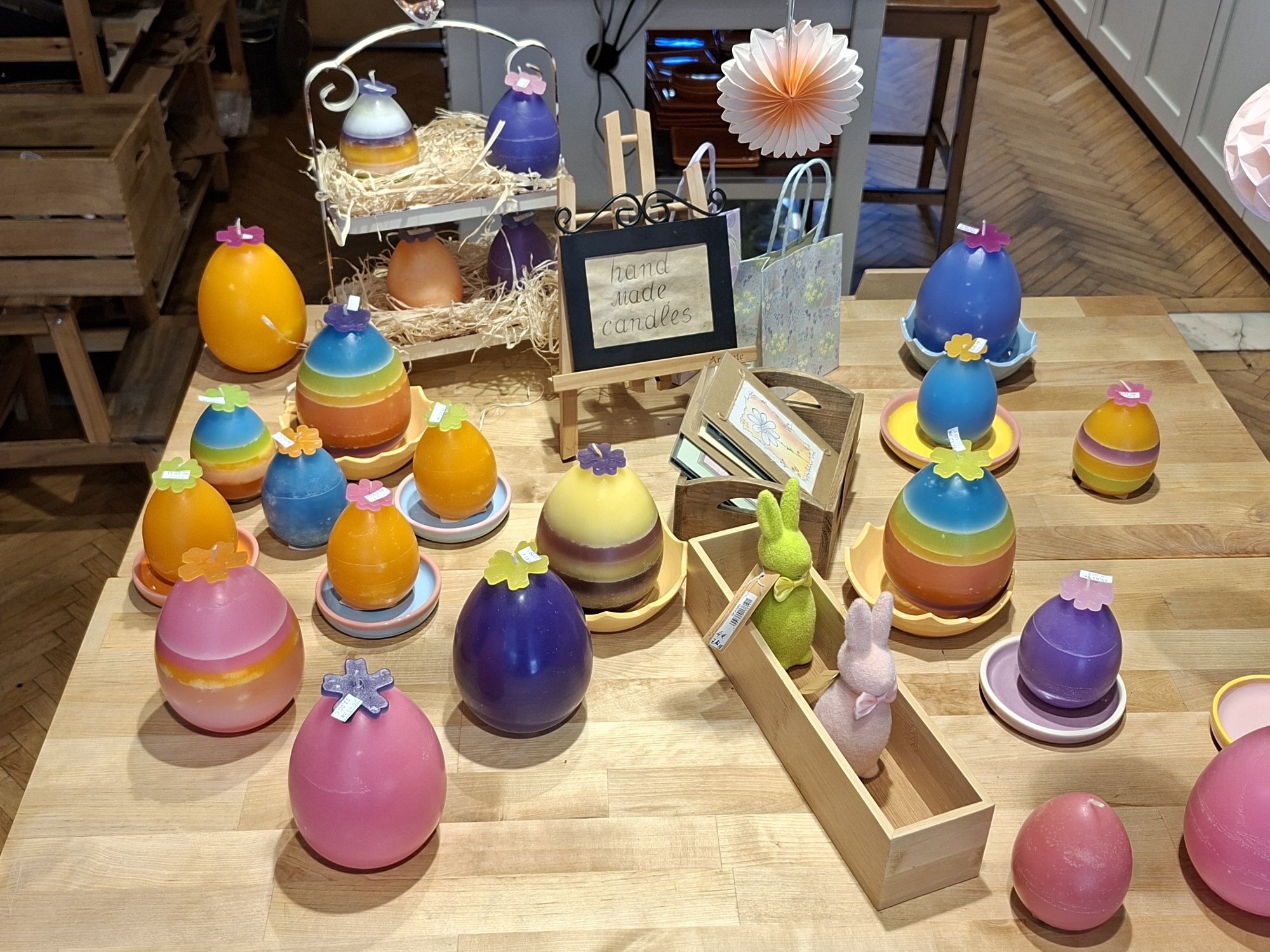 The image shows a display of handmade egg-shaped candles and Easter decorations on a wooden table. The candles are of various colors, including yellow, orange, pink, purple, and blue, often with a decorative flower on top. Some candles are placed on small dishes or plates. Behind the candles, there's a sign that reads "hand made candles" on a small easel. There's a wooden crate with cards and a wooden box with a green and a pink bunny. In the background are parts of other furniture and decorations, including a hanging paper lantern and a wooden crate structure. The location is likely a shop or market stall, but the image doesn't explicitly confirm that it is in Sofia, Bulgaria. The lighting is bright and the overall scene looks festive and intended for a sale display.