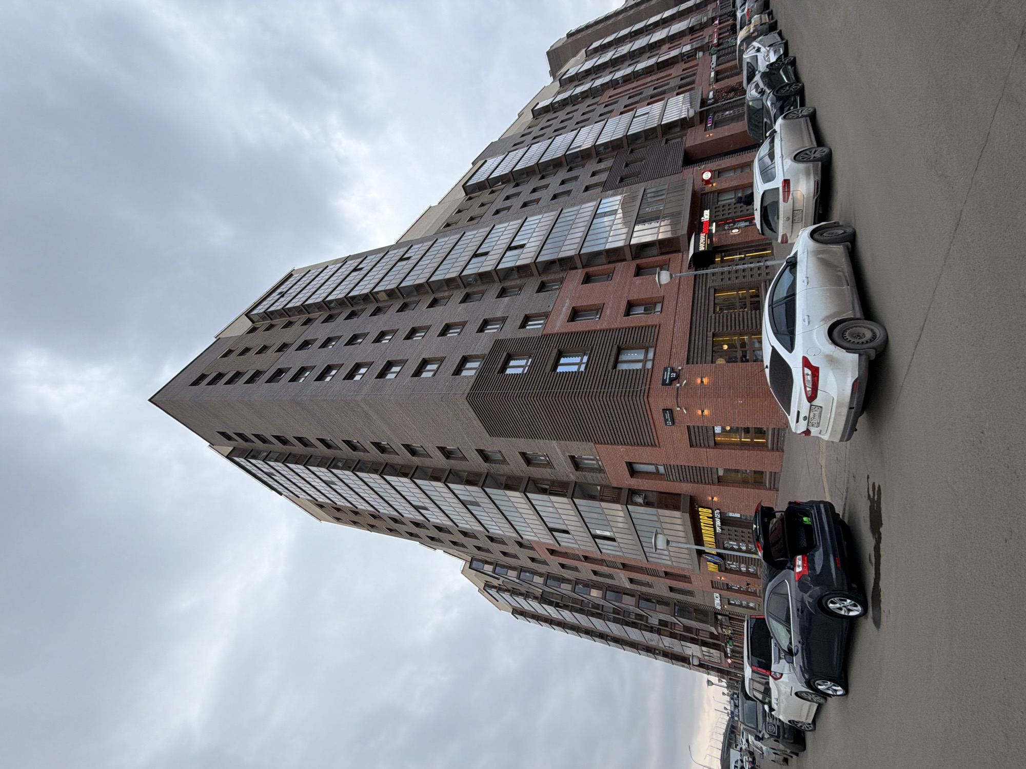 The image depicts a tall, modern brick building in what appears to be Krasnoyarsk, Russia. The building has numerous windows and a stepped design towards the top. Several cars are parked along the adjacent street. The sky is overcast. The presence of a street sign with Cyrillic text and possibly the building's architecture hints at the Russian location. There are no people visible in the frame. The building dominates the scene.
