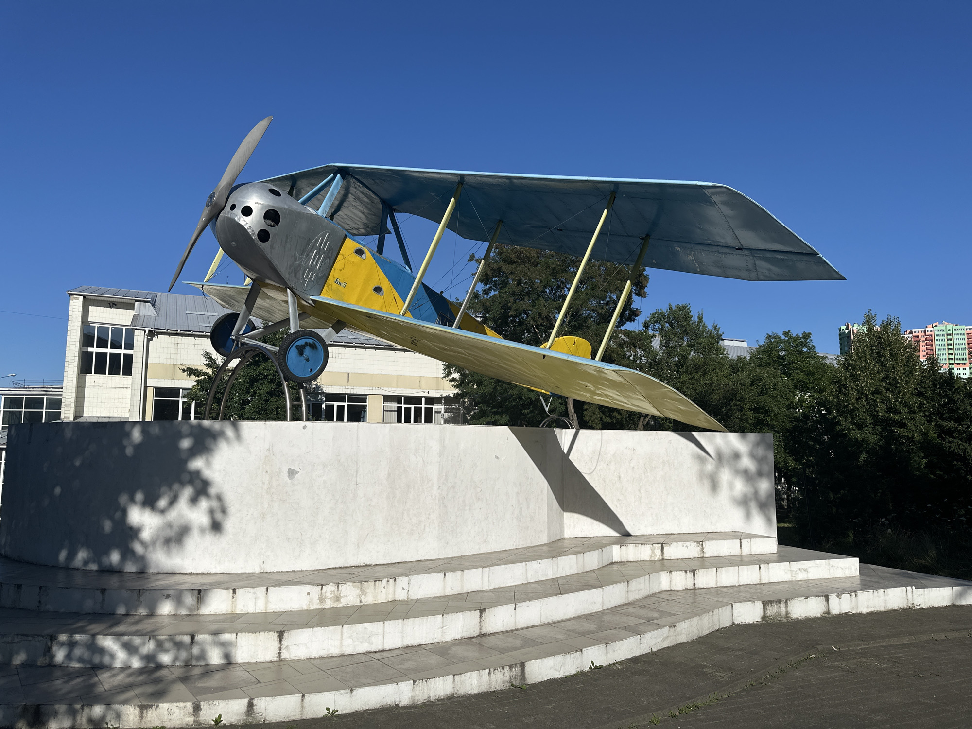 The image shows a biplane sculpture on a monument in Kyiv, Ukraine. The biplane is painted yellow and blue, with a silver fuselage and propeller. It is mounted on a white, stepped concrete base. Behind the monument, there is a building with a glass facade, a line of trees, and a colorful apartment building in the distance. The sky is clear and blue, and the sun is casting shadows on the monument. The biplane appears to be a historical replica or a decorative art piece, possibly commemorating aviation history or a local landmark. The overall setting is an outdoor public space, likely a park or a square, given the monument and surrounding buildings.