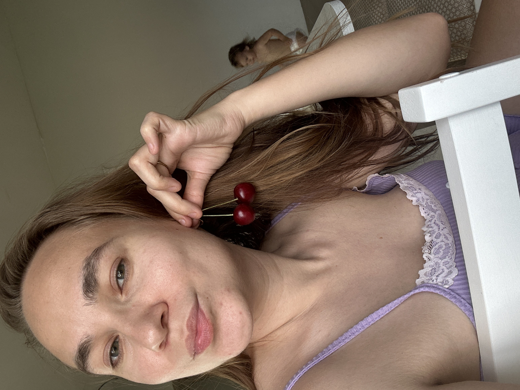 This selfie-style portrait features a young woman with light brown hair and fair skin looking directly at the camera with a soft, content expression. She is wearing a light purple ribbed tank top with white lace trim. Her right hand is raised to her ear, where she playfully holds two vibrant red cherries by their stems, making them appear as temporary earrings.
In the blurred background, to the upper right, a small child, possibly a toddler, is visible, lying or resting on a light-colored surface and wearing a white diaper or undergarment. A segment of a white, angular piece of furniture, likely a chair or bed frame, is also present on the right side of the frame. The setting appears to be an indoor home environment, possibly in Kharnavy, Russia, with a plain, light-colored wall in the background. The lighting is soft and natural, suggesting it is daytime. No text is visible in the image. The scene conveys a relaxed and informal atmosphere.