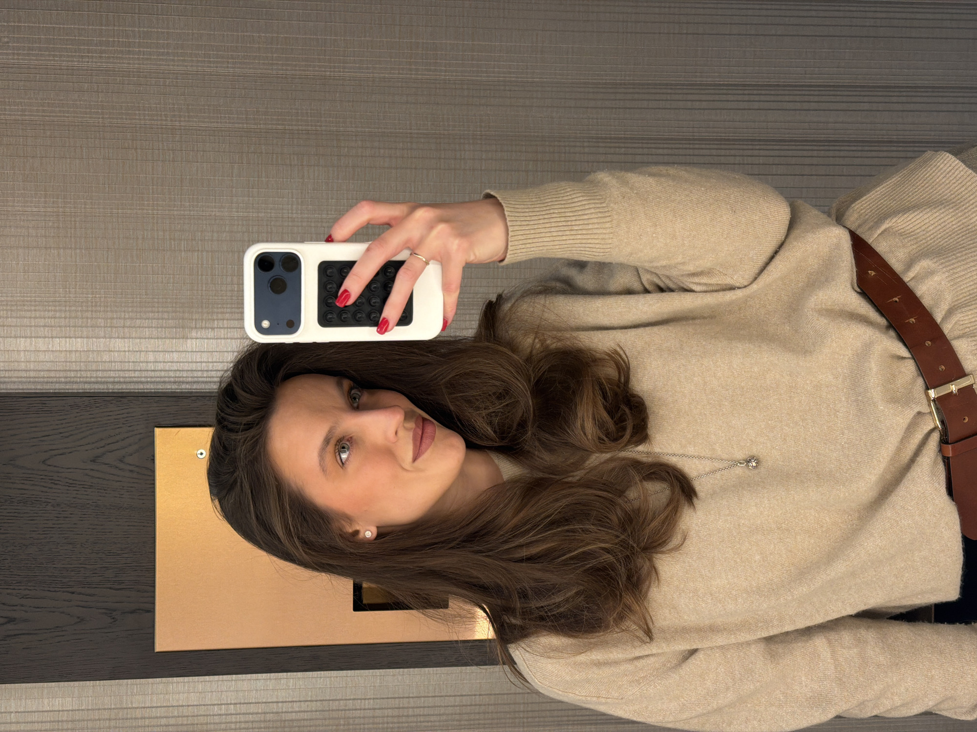 A woman with long brown hair is taking a selfie with a white phone case with a dark blue and black grip. She has red nail polish and is wearing a beige sweater and a brown belt with a gold buckle. The background is a wall with a wooden panel and striped wallpaper. The location is not identifiable from this image.