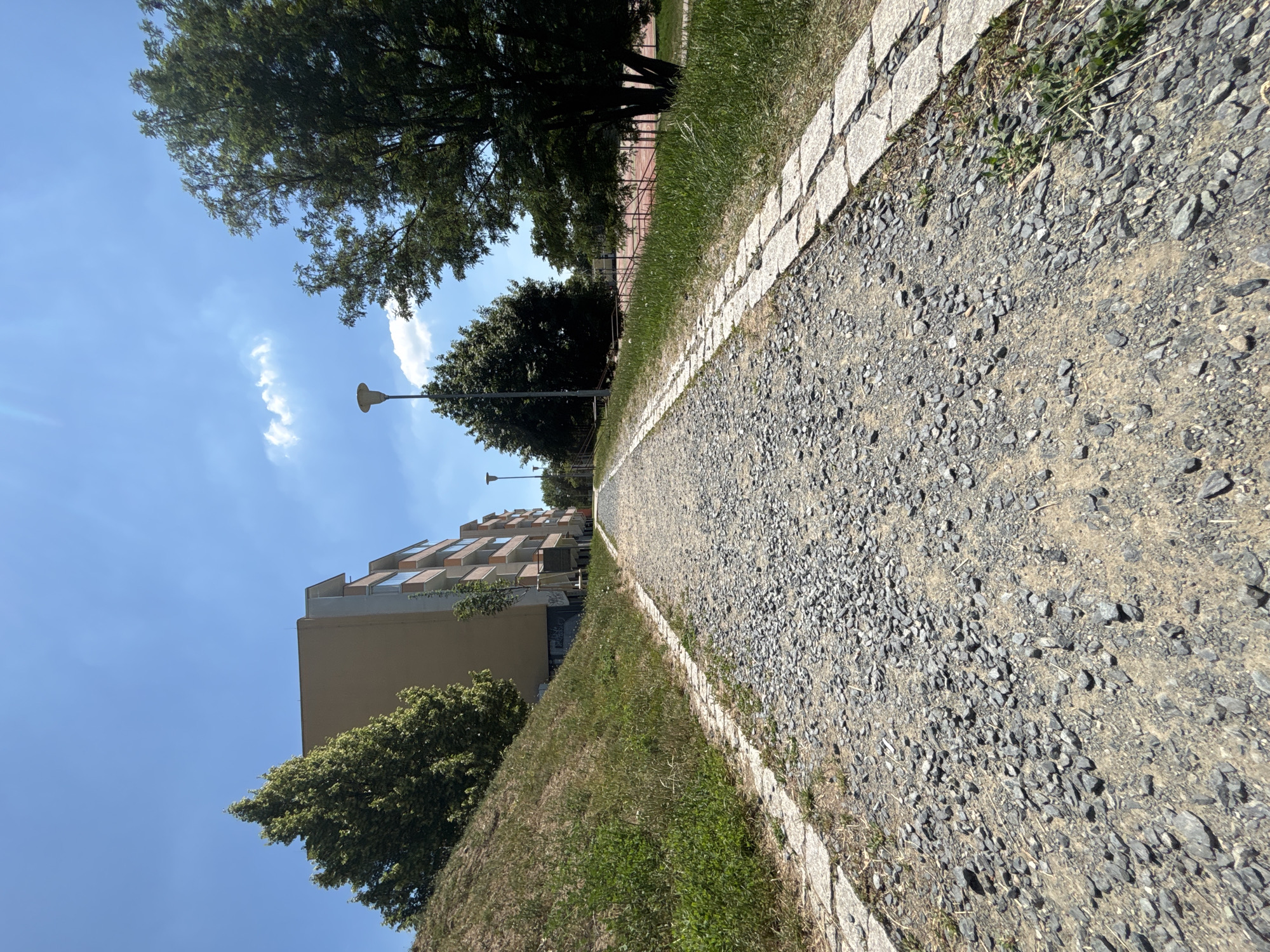 An eye-level, low-angle outdoor photograph captures a gravel path leading into the distance under a clear blue sky with scattered white clouds. The path is lined on both sides by stone pavers and grass. On the left, a grassy slope rises up to a light brown building with multiple balconies and windows. Trees are scattered throughout the scene, with a large, dark green tree dominating the right side of the image. In the middle ground, a street lamp stands near another building. Beyond the trees on the right, a red sports court with a net is visible. The overall impression is a sunny day in a residential or park-like urban setting.