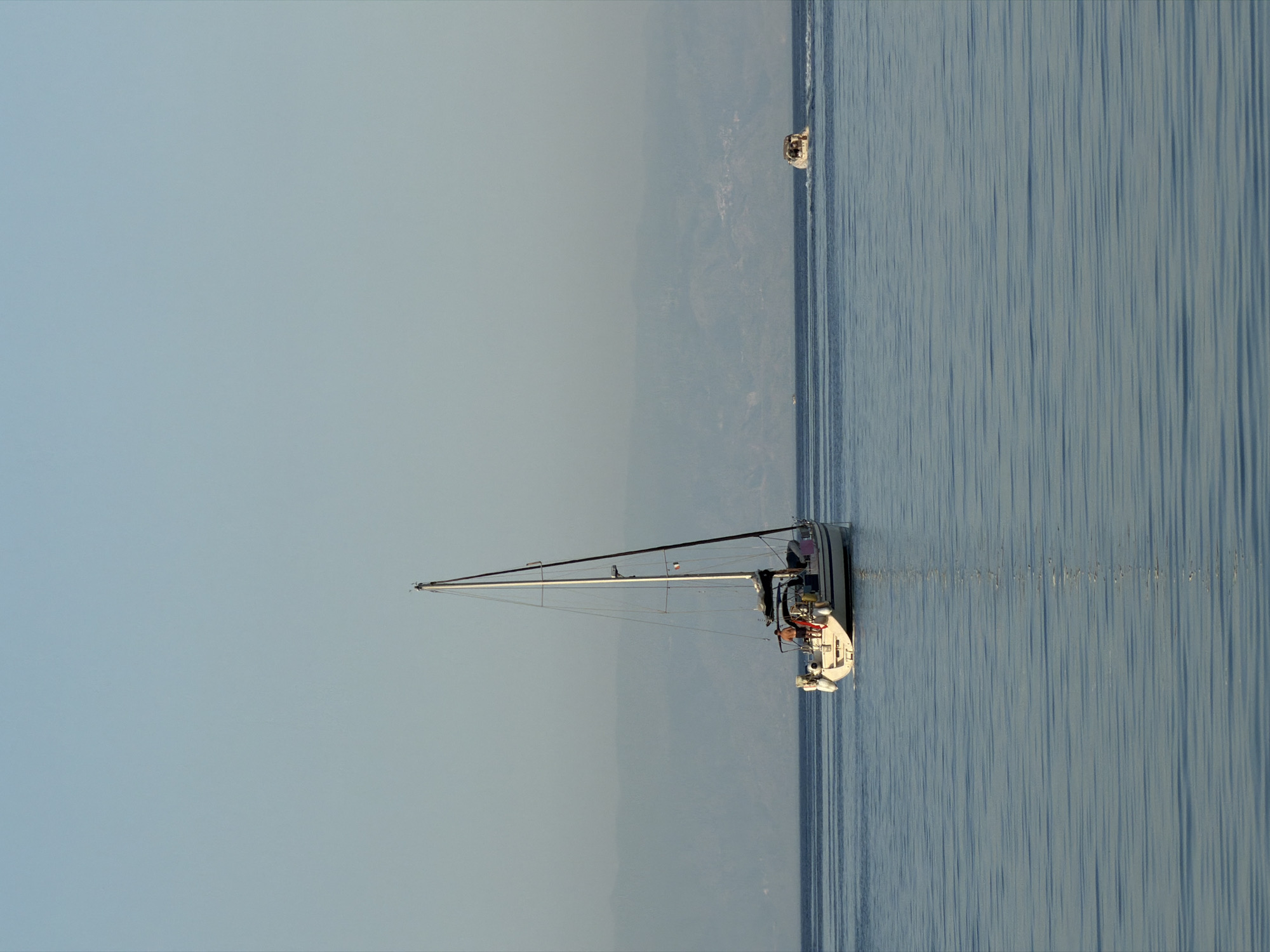 The image depicts a serene maritime scene. A white sailboat with its mast raised dominates the right side of the frame, floating on calm water. In the background, a distant boat is visible. The sky and the sea meet at the horizon, with a subtle coastline suggesting the location. The color palette is predominantly blue and white, reflecting the sky and water. There is no evidence to support the claim the image is in the city of Chianchitta, Italy.