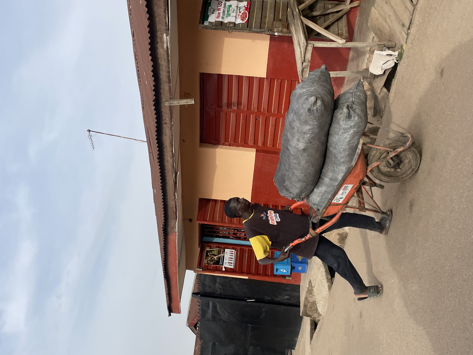 An outdoor scene in Jos, Nigeria, depicts a man pushing a wheelbarrow loaded with two large grey sacks. The man, seen from the waist up, wears a dark t-shirt with yellow short sleeves and dark trousers, along with open-toed sandals. Text on his shirt reads "Rookies CEREAL". He is pushing a bright orange or red metal wheelbarrow, which has a single wheel and appears to be heavily laden. Partially visible text on the wheelbarrow indicates it was "MANUFACTURED IN KANO".
In the background, a building with an orange and red painted facade features a corrugated metal roof. A red door or shutter is visible on the building. Above the roof, against an overcast sky, a simple antenna with multiple elements is present. Below the main building, a darker structure or wall is partially visible. Near the building's entrance, two blue plastic crates are stacked, and a poster on the wall shows people and indistinct text. Another advertisement, likely for a food product, displays "NOW N 120" and "Max" several times, along with "16 cubes inside". The ground is a light concrete or paved surface.
The setting suggests a typical day in a bustling local area, with the man engaged in transporting goods. The overcast sky indicates daytime without direct sunlight.