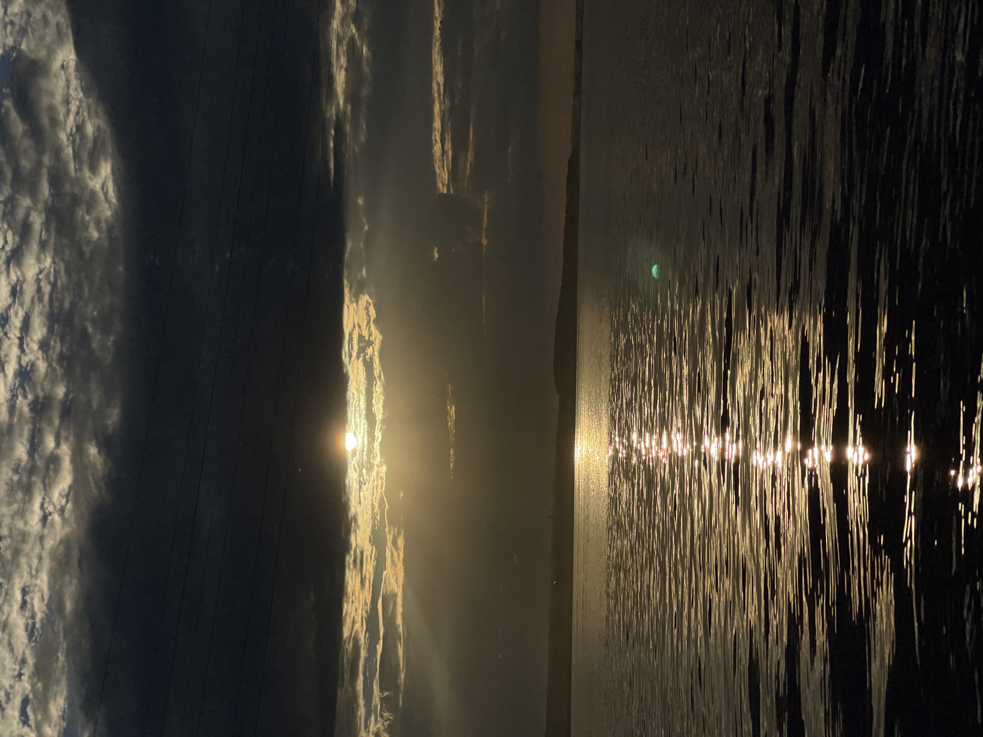 The image displays a dramatic seascape with a sunset or sunrise casting a golden glow over the scene. The sky is filled with a mix of dark, stormy clouds and illuminated, wispy clouds that catch the light. In the foreground, there appears to be a body of water, possibly the ocean or a large lake, with the light from the sun or moon reflecting off its surface in a shimmering, broken pattern. The reflection creates a luminous pathway across the water.
The overall atmosphere is one of intense natural beauty and perhaps a touch of melancholy due to the dark clouds juxtaposed with the bright light. The time of day is clearly indicated as either dawn or dusk by the low angle of the sun and the warm, golden hues. The weather appears to be dynamic, with the presence of both heavy clouds and clear patches allowing sunlight to break through.
There are some vertical lines visible in the upper left portion of the image, which could suggest the presence of power lines or similar structures, though they are not the primary focus. The vastness of the water and sky suggests an open, natural landscape, possibly a coastal area or a large inland body of water. There are no people or animals visible in the frame, emphasizing the solitude of the natural environment. The emotions evoked are awe, wonder, and a sense of the power and beauty of nature. No text is visible in the image.
