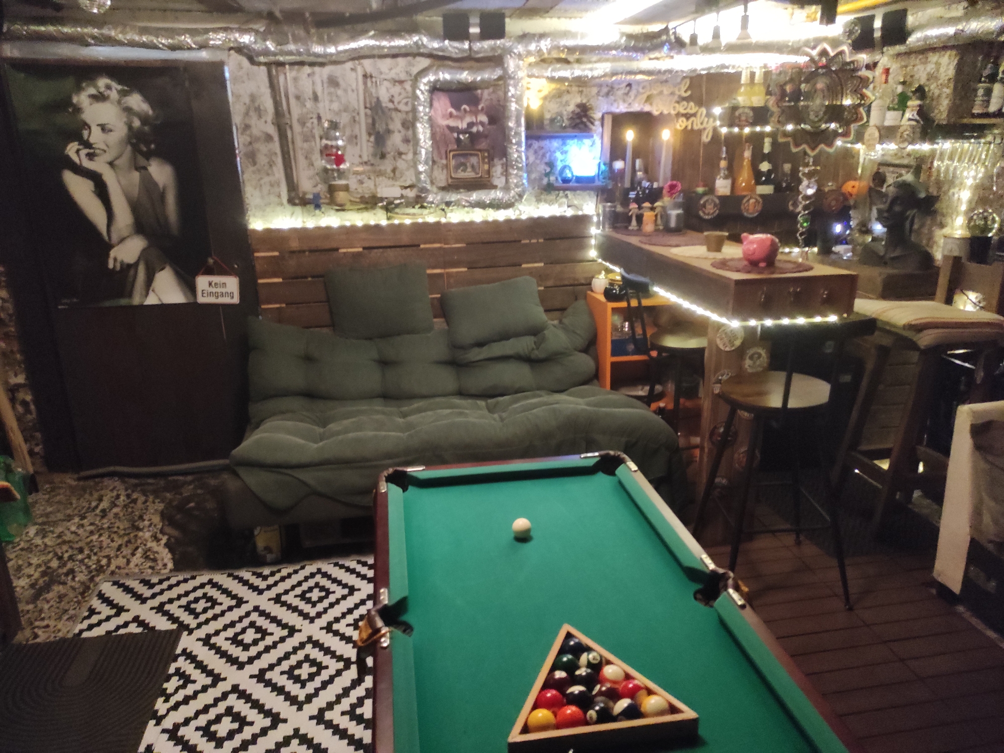 The image depicts an indoor recreational space that appears to be a private bar or lounge. The room is dimly lit, with strings of fairy lights illuminating the walls and ceiling. In the foreground, a green pool table is set up, with a rack of billiard balls and a single cue ball in position. A portion of a patterned rug is visible in the lower left corner.
To the left of the pool table, a dark green futon sofa is arranged against a wooden slatted wall. Above the sofa, a large black and white portrait of Marilyn Monroe is hung. A small sign reading "Kein Eingang" (No Entry) is attached to the lower right of the portrait.
The background features a bar area with various bottles of alcohol displayed on shelves and the bar counter. Decorative items, including what appear to be figurines and a small television, are also visible. The walls are textured, with exposed pipes and a metallic sheen in some areas, suggesting a basement or converted space. The overall atmosphere is informal and designed for leisure. The image is located in Waldbronn, Germany.