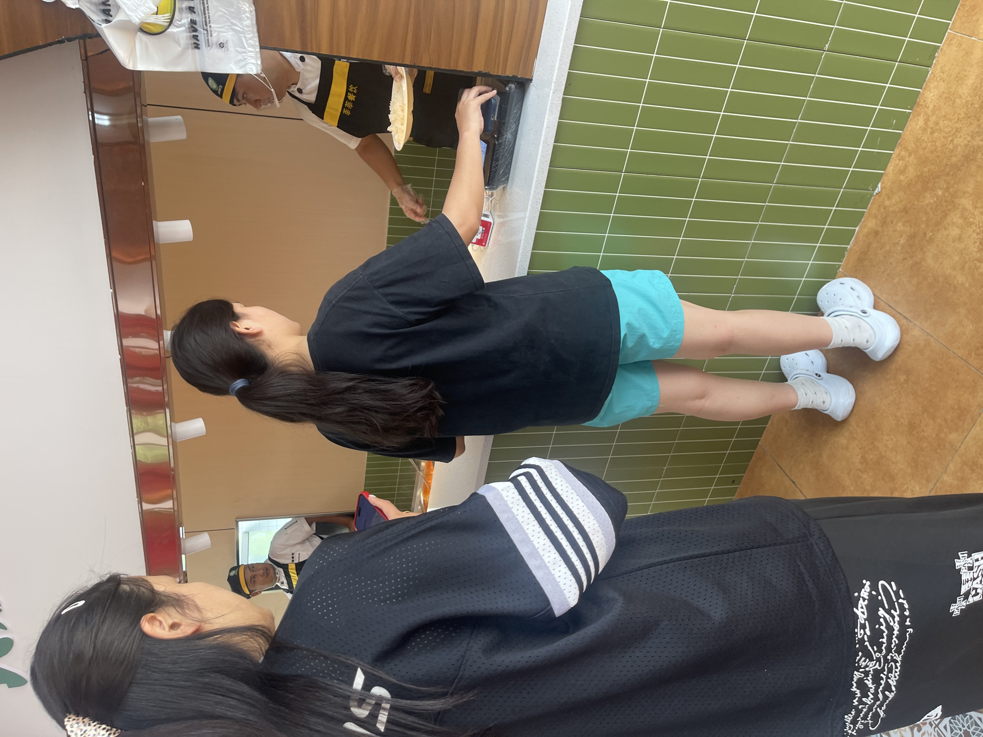 The image depicts an indoor scene in what appears to be a food service establishment. A person wearing a black t-shirt and turquoise shorts stands near a counter. Behind the counter, a food service worker wearing a uniform (white shirt, black apron with writing in Chinese) is preparing food. A second person wearing a black Adidas-branded jacket or sweatshirt stands in the foreground. The floor is partially covered with green tiles, transitioning to a brown-toned surface. Reflections in the surrounding surfaces indicate that the scene is located in Chongqing High-tech Zone, China.