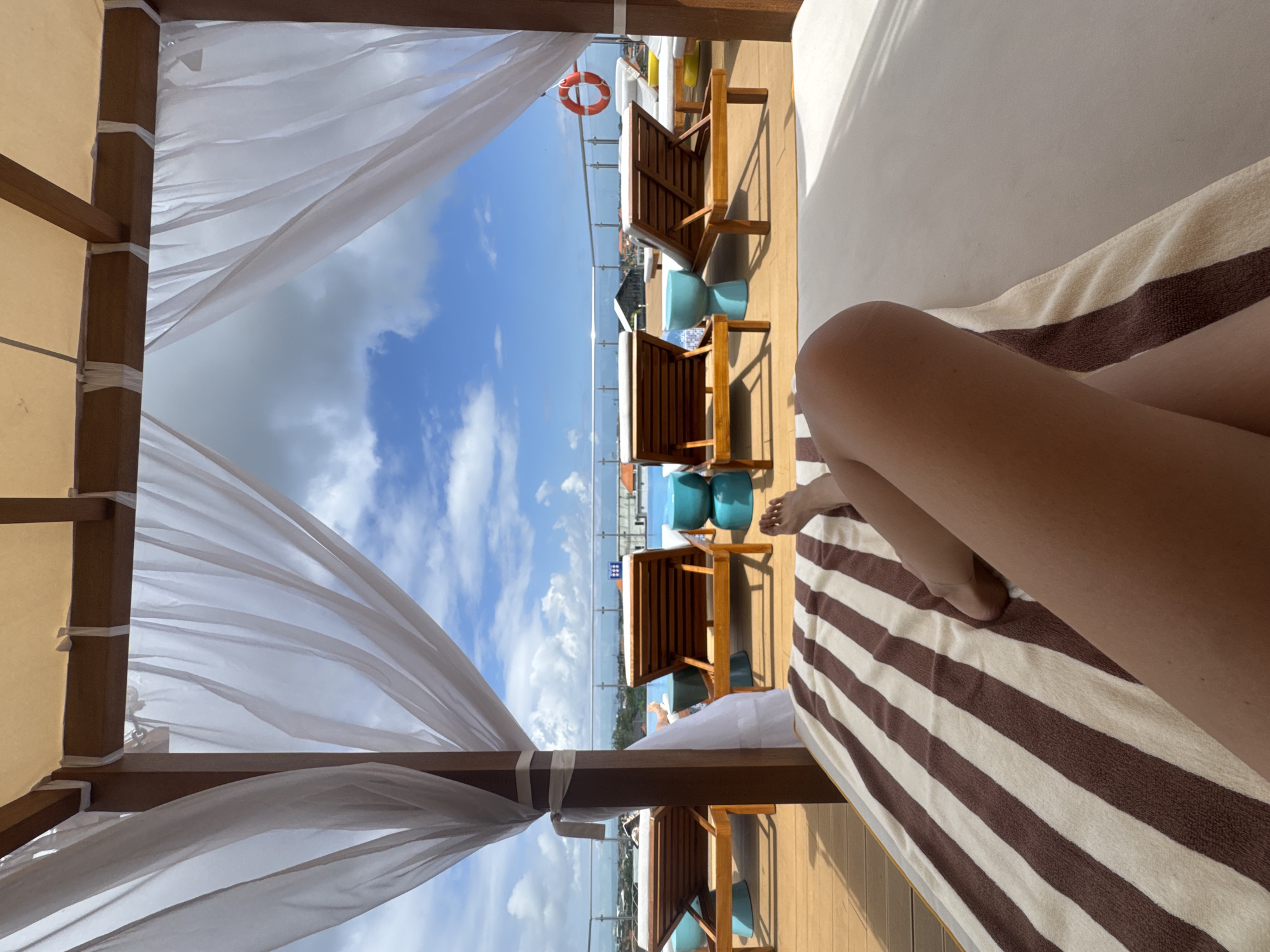 This is a first-person perspective shot from a covered lounge chair on a rooftop deck, looking out towards a swimming pool and the city of North Kuta, Indonesia. The lounge chair has a brown and white striped towel. In the foreground, a person's legs are visible, resting on the towel. Beyond the lounge chair are several empty wooden lounge chairs with white cushions, some with turquoise cylindrical stools beside them. A pool is visible in the middle ground, with a railing and then the cityscape beyond. The sky is blue with scattered white clouds. A life preserver ring is hanging on the railing to the right. The covered area has wooden beams and light white sheer curtains.
