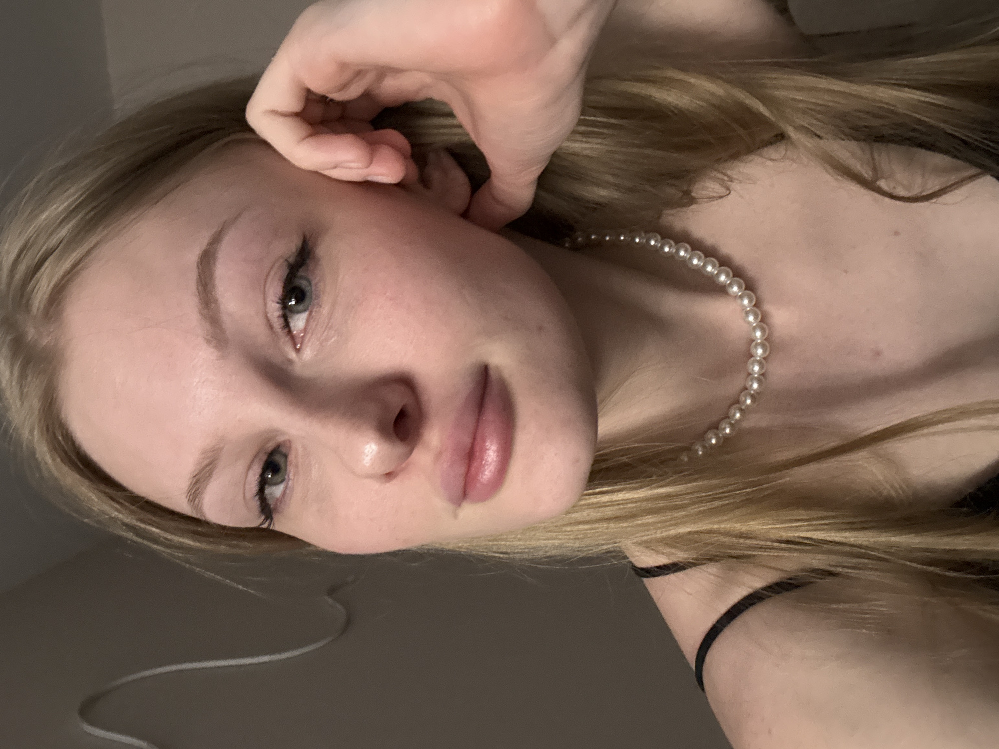 A young blonde woman with light skin and blue-green eyes is shown in a close-up portrait. She has on a black slip dress and a pearl necklace. Her left hand is resting on her cheek. The background is a plain, light-colored wall with a subtle decorative element. The image does not contain any information that would suggest a specific location in Perm, Russia, or any other city.