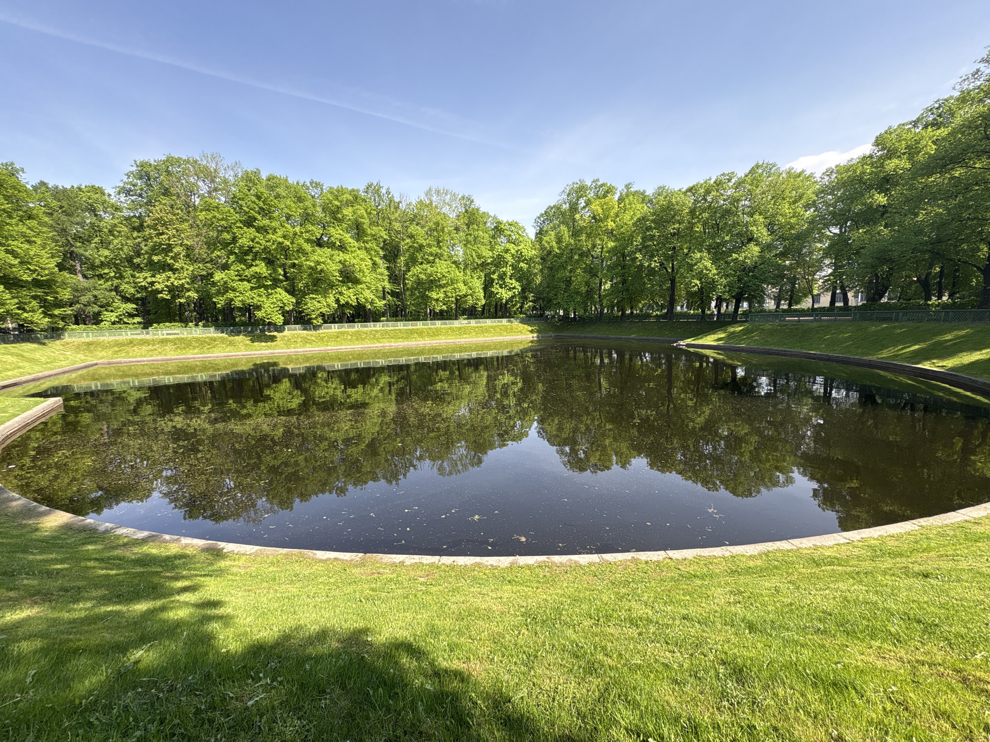 The image depicts a circular pond in a park, likely located in Saint Petersburg, Russia, given the architectural style of the surrounding fencing and the typical park layout. The pond is surrounded by neatly manicured green lawns and a low concrete retaining wall. The water surface reflects the surrounding trees, which are lush and green, suggesting a summer or spring season. A blue sky with faint clouds is visible above the trees. There are no people visible in the scene.