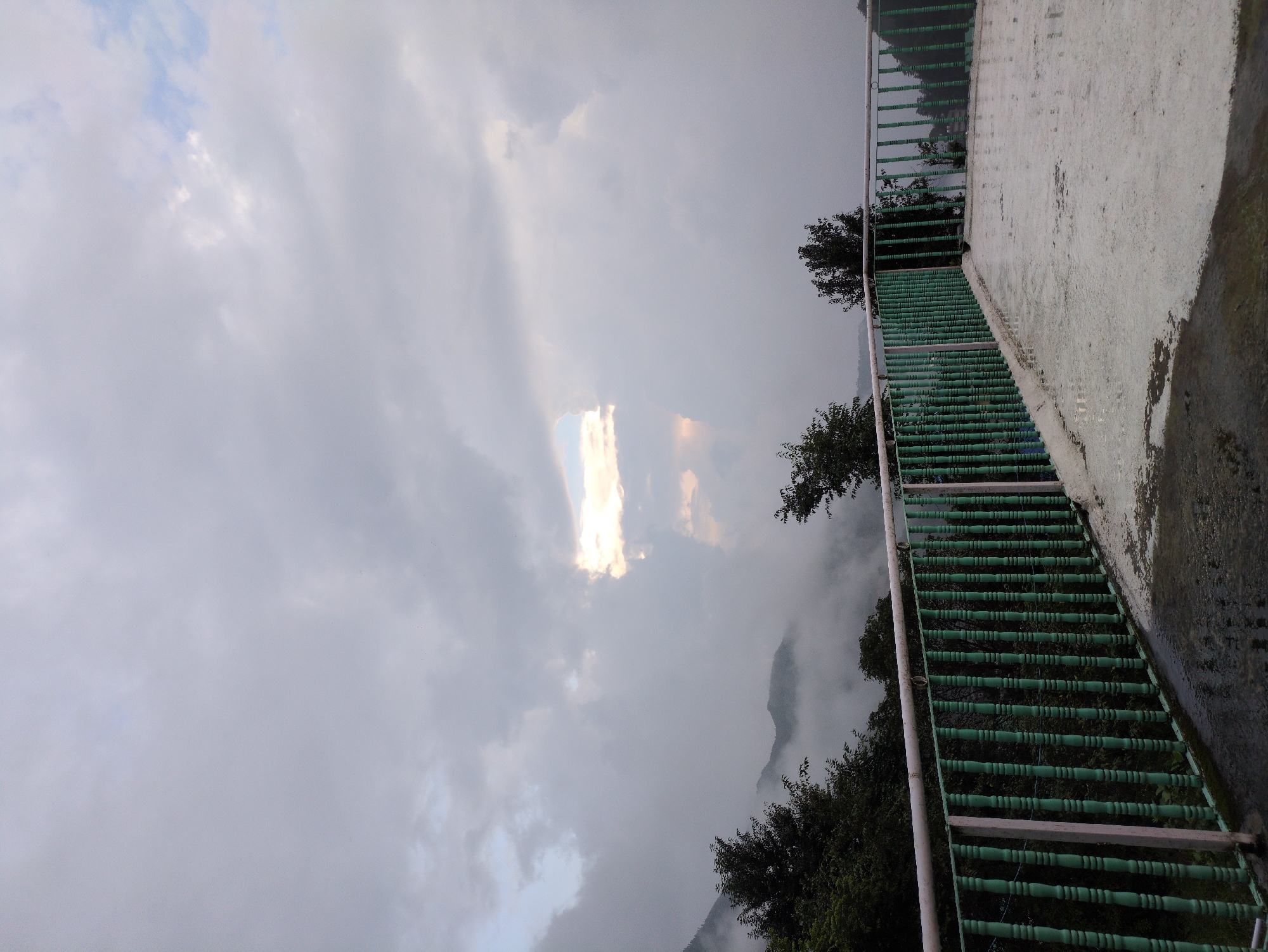The image depicts a view from a rooftop in Bhagsu, India, on a cloudy day. In the foreground, a concrete floor with puddles suggests recent rain. A green railing with white posts runs along the edge of the rooftop, separating it from a steep, forested slope below. The slope is covered with dense trees, some of which are silhouetted against the mist. In the background, mountains are visible, partially obscured by clouds and fog, with a patch of sunlight breaking through the overcast sky, illuminating a portion of the clouds and the distant landscape. The overall atmosphere is serene and slightly melancholic, typical of a monsoon or cloudy season in the mountains.