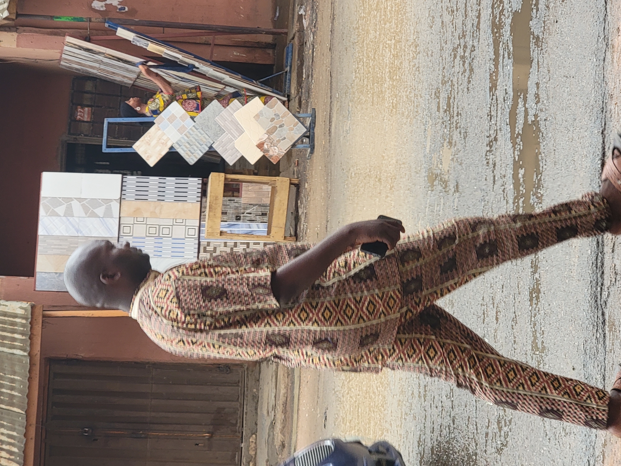 The scene captures a man walking on a street in Maiburiji, Nigeria. He is wearing patterned brown clothing and holding a mobile phone. Behind him, there is a display of tiles outside a building. The street is wet, possibly from recent rain. There is another person in the background near the tiles.