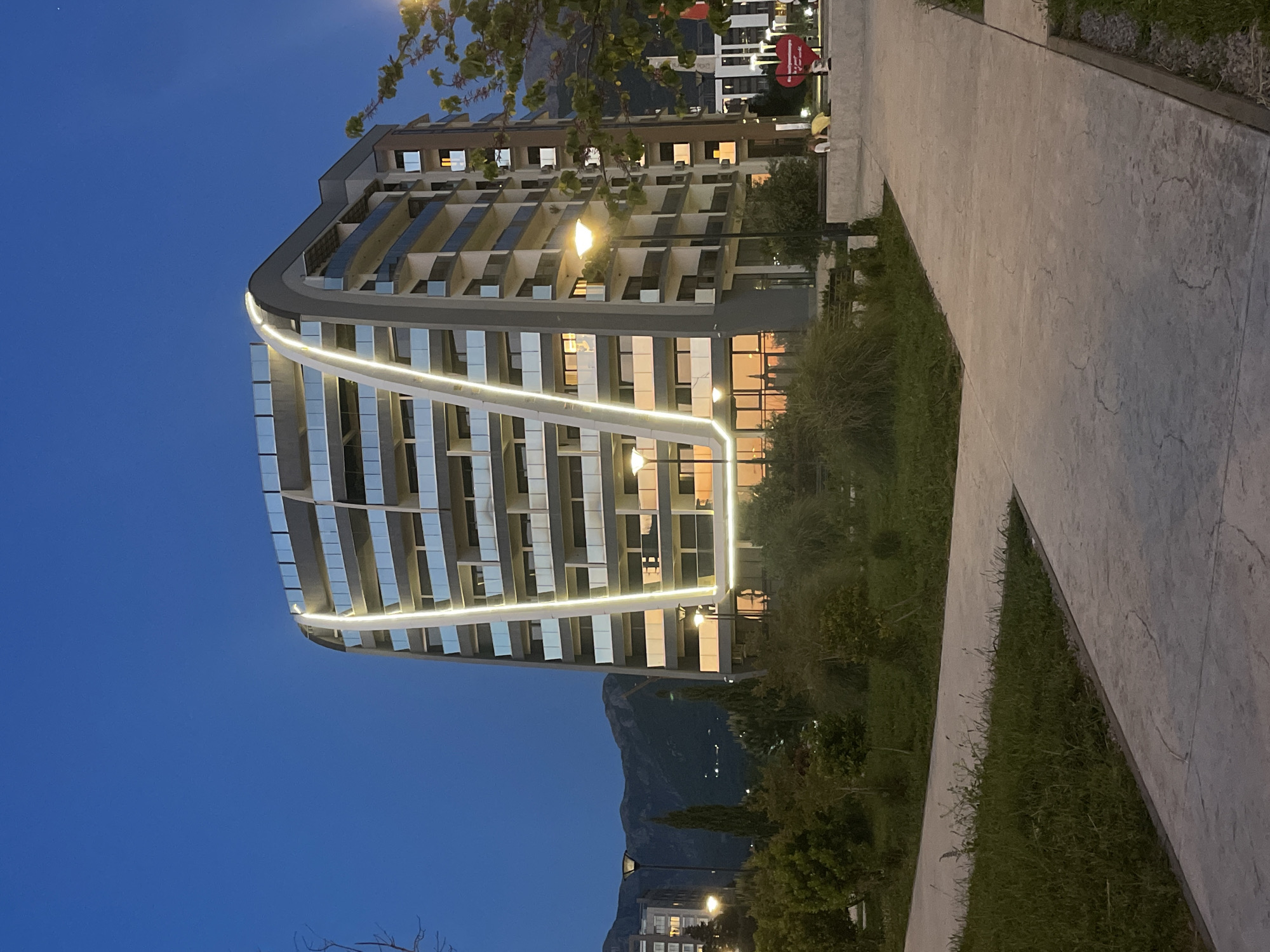 The image displays a modern multi-story building at dusk or night in Bar, Montenegro. The building, oriented vertically, features a distinctive curved facade with numerous balconies and reflective glass windows. A prominent, bright white LED light strip accentuates the building's contemporary design, following its graceful curve from top to bottom. Warm interior lights glow from many of the windows, indicating occupancy and adding to the evening ambiance.
In the foreground, a light-colored, paved pathway with textured patterns runs diagonally, separating grassy areas from the viewer. Scattered trees and shrubs are visible in the surrounding landscape. Looming mountains define the distant background against a deep blue twilight sky, indicating a clear evening.
Near the right side of the building, in what appears to be a public space or promenade, two indistinct figures are visible. They stand near a prominent red, heart-shaped decorative object, which has white text "LOVE" emblazoned across it. Other background structures and streetlights are also faintly visible. The scene portrays a calm, illuminated evening in a modern setting, blending contemporary architecture with natural scenery.