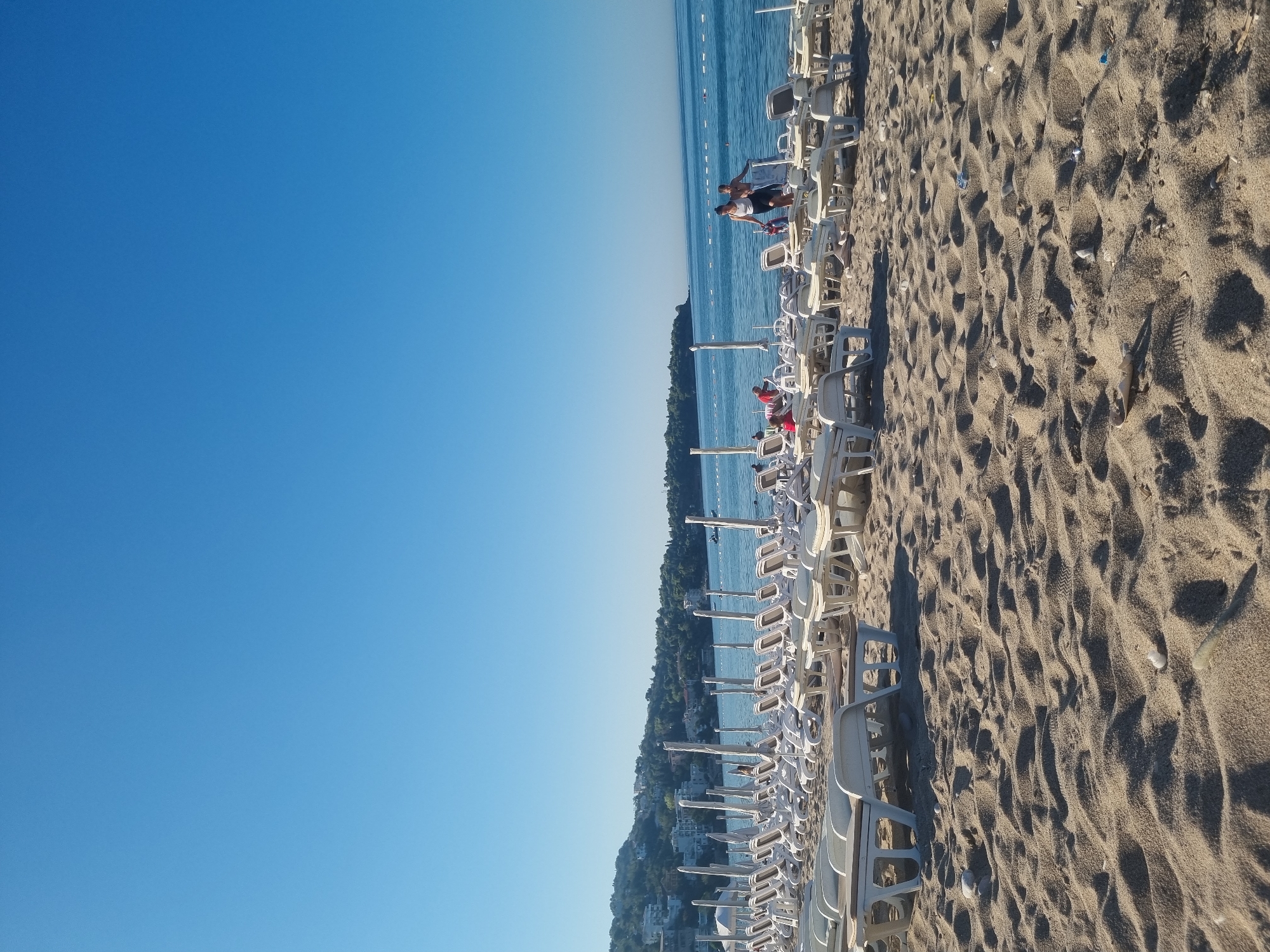 The image depicts a sunny day on a sandy beach in Sutomore, Montenegro. The foreground is dominated by textured sand, with ripples and scattered small stones. Behind the sand, rows of empty white lounge chairs with integrated tables are neatly arranged, facing the sea. Some chairs are slightly angled. In the middle ground, a few people are visible. Two individuals, one in a dark skirt and white top and the other in a dark top and skirt, walk near the water's edge, carrying a red bag. Another person in red sits on a lounge chair further back. The sea is calm and blue, with a thin white buoy line stretching across it. In the background, a hillside covered with green trees and white buildings forms a picturesque backdrop to the coastline. The sky is a clear, bright blue.