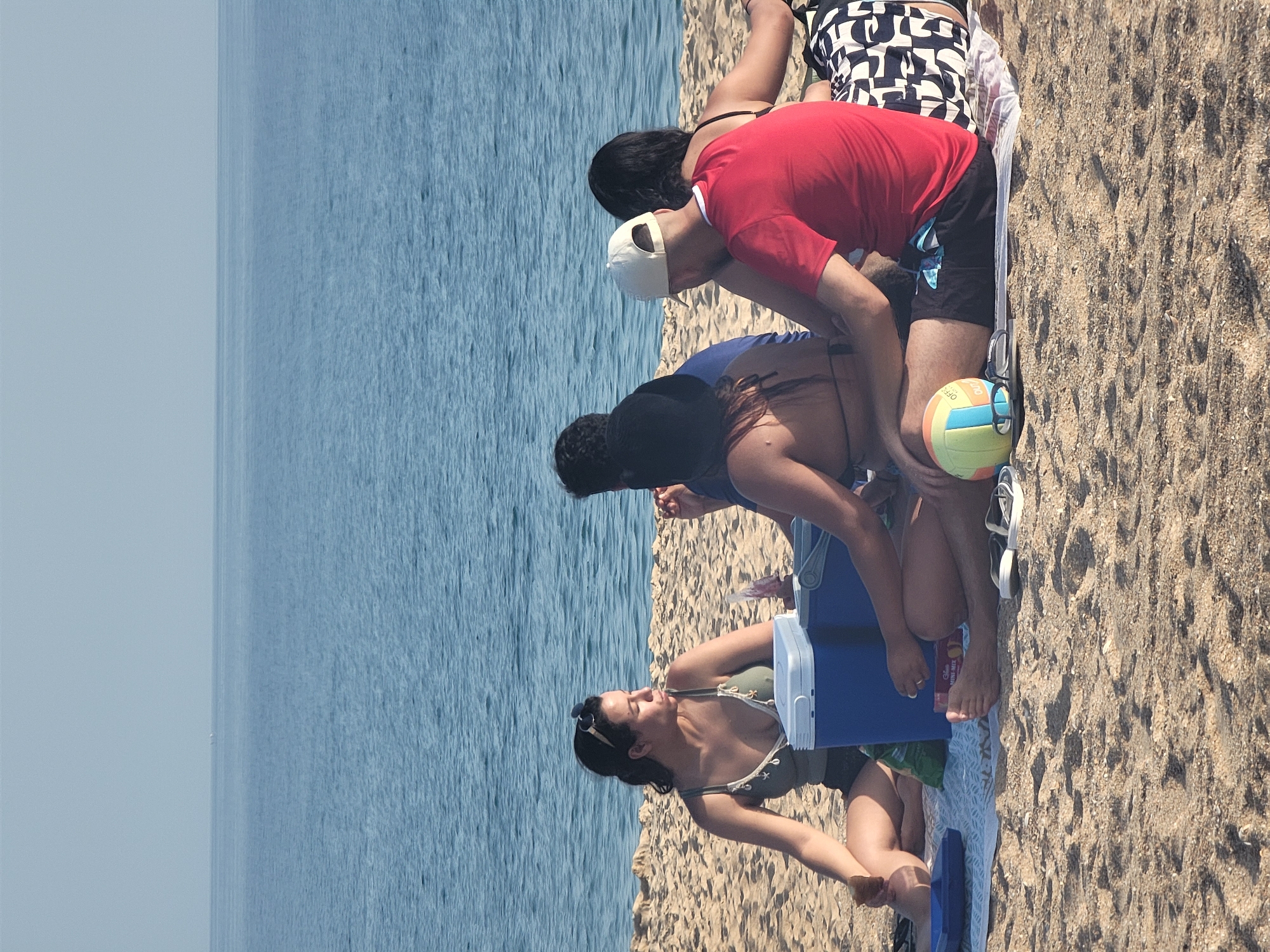 A group of four people are having a picnic on a sandy beach in Quarteira, Portugal, with the ocean in the background. The woman on the far left is wearing a green top and dark shorts and is looking to her right. Next to her is a blue cooler. In the center, two people are seated with their backs to the camera. The person on the left is wearing a blue shirt and a black hat, and the person on the right is wearing a red t-shirt and a white baseball cap. The woman on the far right is wearing a black bikini top and patterned shorts and is facing away from the camera. A volleyball is on the sand in front of the group, and a pair of sunglasses is next to it. The sky is clear and the water is calm.