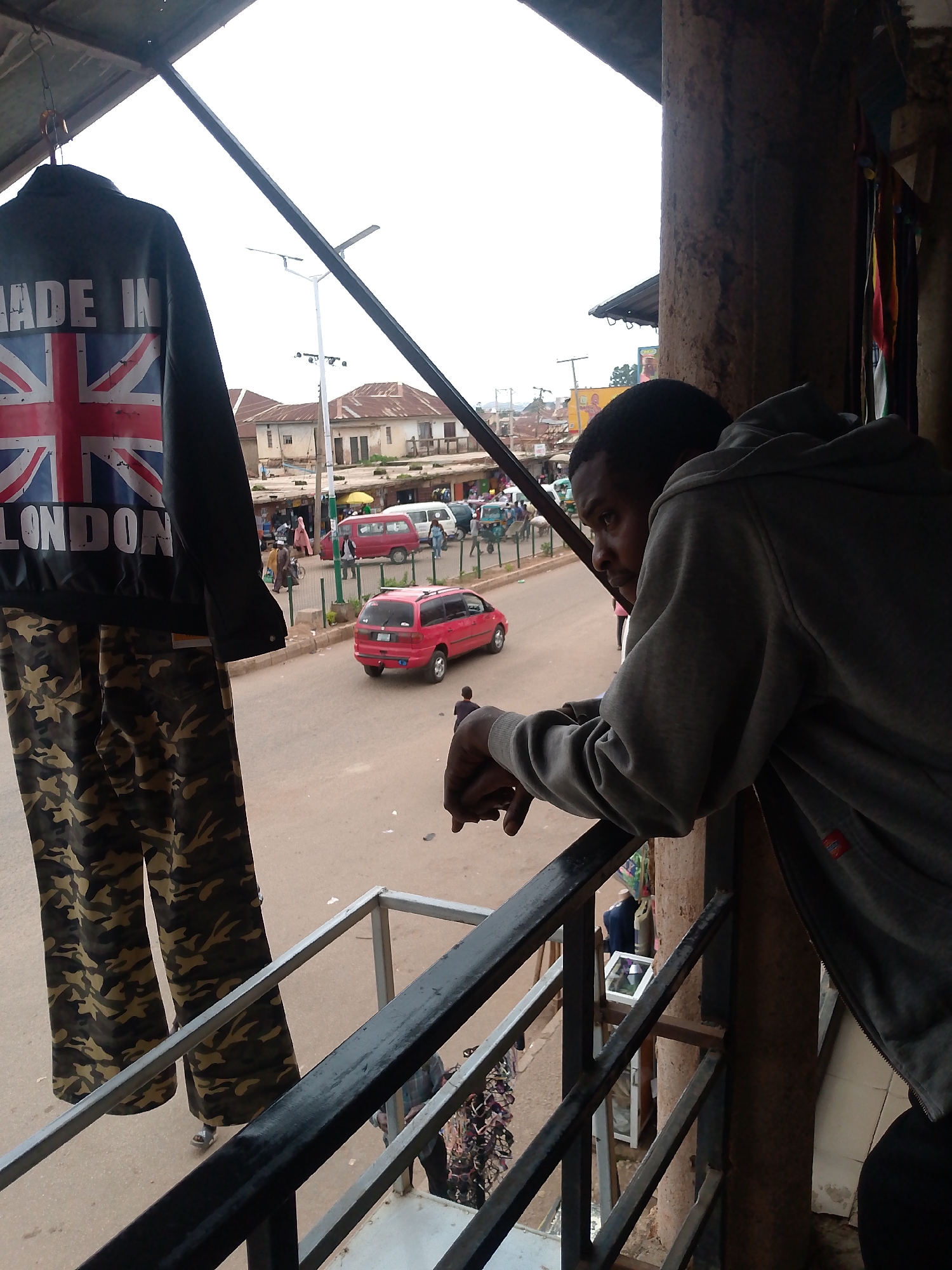 A young man in a grey hooded sweatshirt leans over a railing, looking down at a street scene in Bukuru, Nigeria. The street below is busy with traffic, including red cars and vans, and pedestrians. Buildings line the street, some appearing to be shops. On a railing to the left, a jacket with the Union Jack flag and the words "MADE IN LONDON" is displayed alongside camouflage pants. The overall impression is one of an active urban environment in Nigeria.