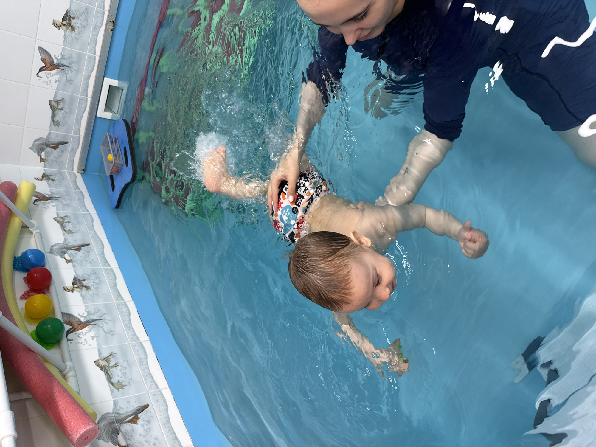 An overhead shot captures an adult and a young child engaging in a swimming activity within an indoor pool in Rusanovo, Russia. The adult, wearing a dark blue long-sleeved swim shirt, is positioned on the right, gently holding and guiding the child. The child, a toddler with light brown hair and wearing colorful patterned swim trunks, is lying on their back in the water, with arms slightly outstretched and legs creating small splashes. The adult looks down at the child with a focused and caring expression, while the child appears calm and content, with eyes slightly closed or looking up.
The pool's edge, visible on the left side of the frame, is lined with white tiles adorned with various bird illustrations, including ducks and geese. Alongside the tiled edge, several swimming aids such as pink, yellow, and green pool noodles, and colorful plastic balls (blue, red, yellow, green) are neatly stored. A blue floating board with a gray basket containing a yellow ball is also visible near a white skimmer or filter vent on the pool wall. The water is clear blue, with surface ripples and reflections indicative of an indoor setting with overhead lighting. No discernible text is visible on clothing or objects. The scene suggests a swimming lesson or playful aquatic interaction between the two individuals.