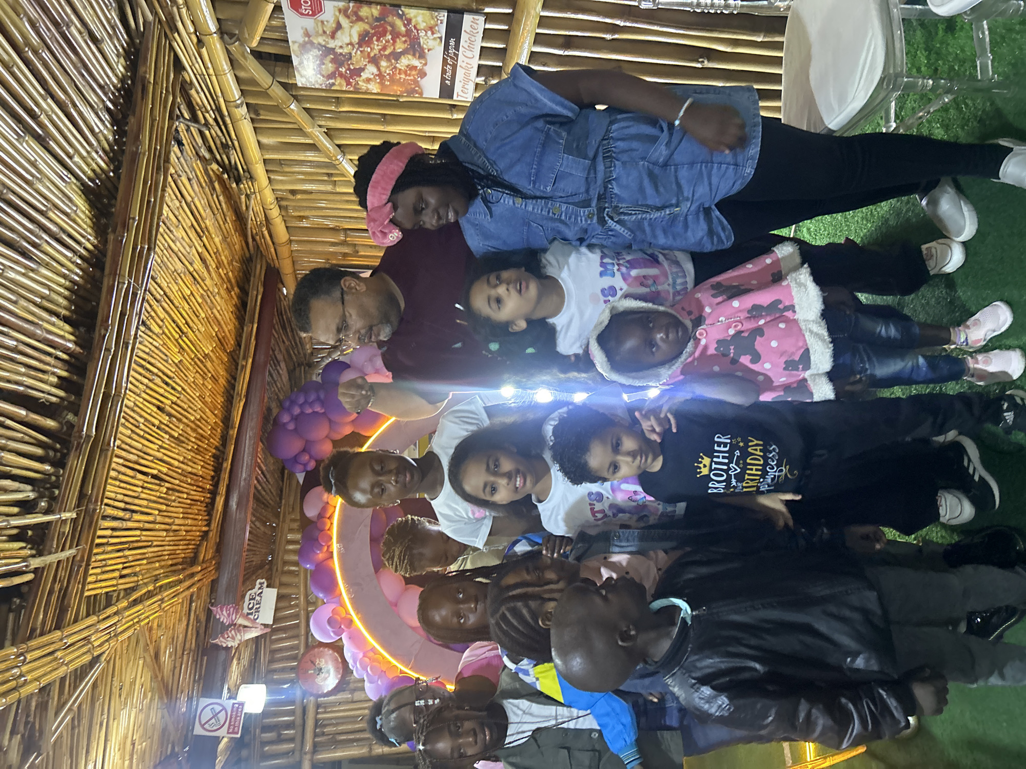 In a brightly lit indoor setting with a thatched bamboo ceiling, a group of approximately ten people, predominantly children and some adults, are gathered. The children appear to be the focus of the event, possibly a birthday celebration, indicated by balloons and a lit arch in the background. The ambiance is festive and casual. The individuals are dressed in everyday clothing. The scene suggests a community gathering or family celebration.