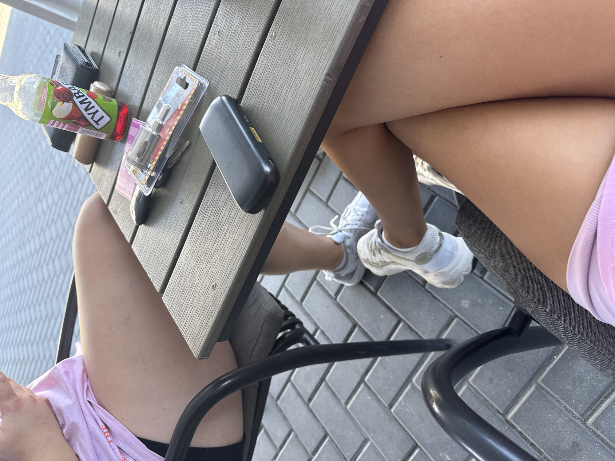 The image shows a close-up view of two individuals' legs and thighs seated at a grey, slatted outdoor table. The table has several items on it, including a bottle of Tymb a beverage with a label featuring apples and cherries, a black wallet, a brown cylindrical object, car keys, and a black rectangular electronic device. The ground beneath the table is paved with grey brickwork. One individual is wearing a pink t-shirt and black shorts, while the other is wearing white sneakers and light-colored shorts. The scene appears to be outdoors, possibly a cafe or restaurant seating area, and is implied to be in Comrat, Moldova.