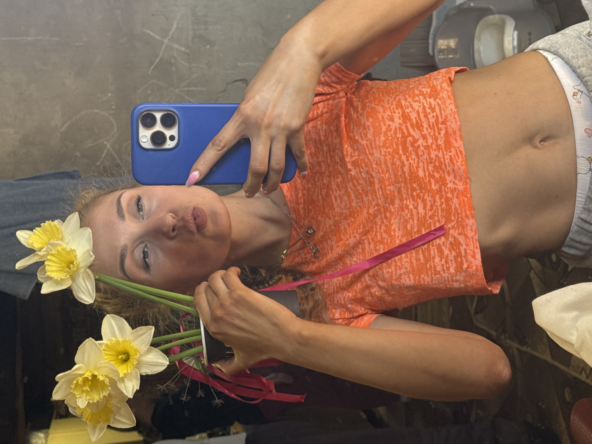 A woman is holding a blue smartphone in her right hand, taking a selfie. She is wearing an orange patterned crop top and grey sweatpants, revealing her midriff. In her left hand, she holds a bouquet of yellow and white daffodils tied with pink ribbons. The background is blurry but appears to be an indoor space with neutral-colored walls and indistinct objects, possibly a bathroom, out of focus behind her. The presence of the daffodils and the casual attire suggest a personal, informal setting. There is no indication in the image that this was taken in Nizhnyaya Chermoda, Russia.