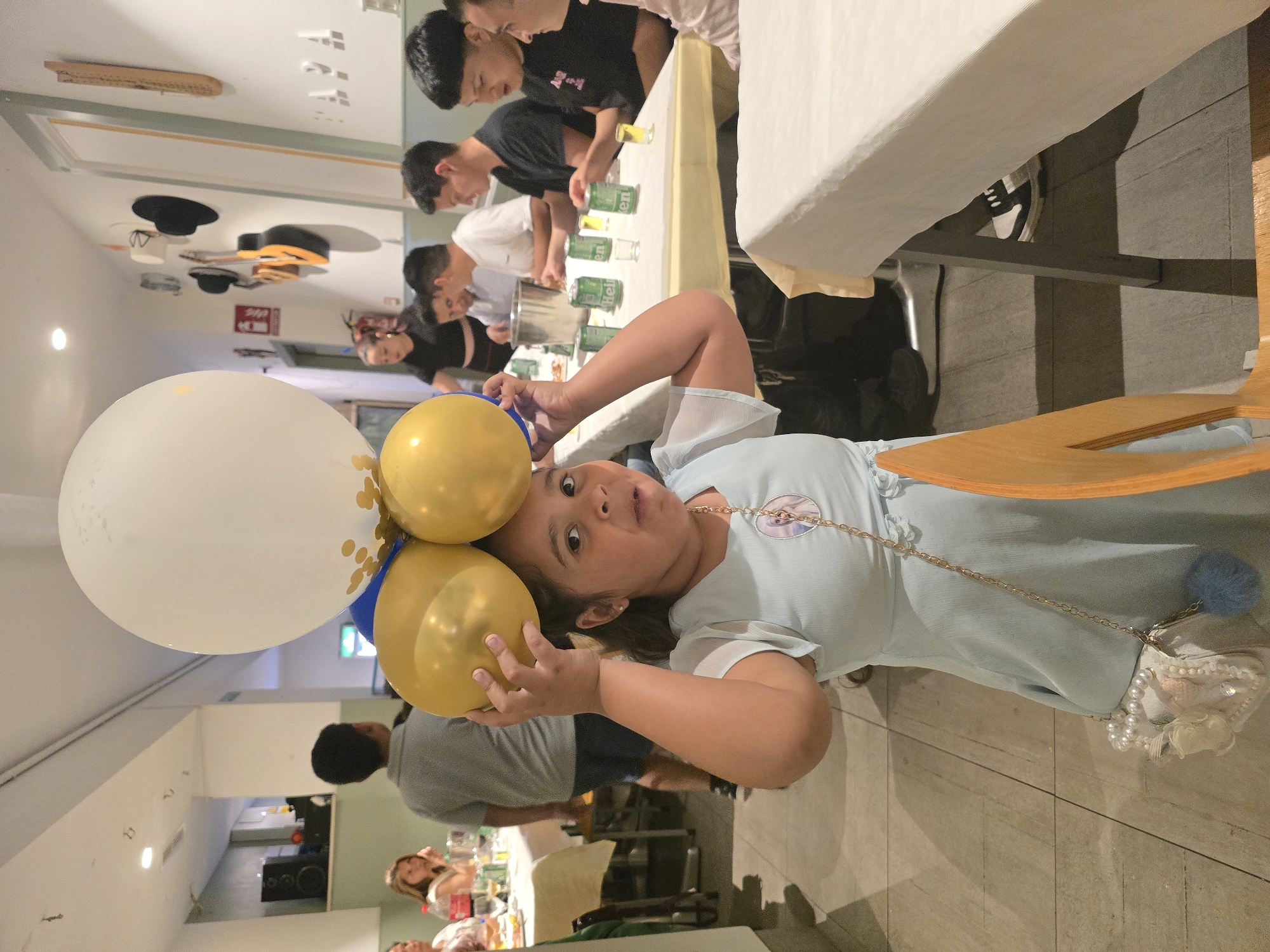 An indoor scene captures a casual gathering or party in L'hospitalet De Llobregat, Spain. In the foreground, a young girl with dark hair, wearing a light blue dress and a small chain bag with a circular character motif, playfully holds three balloons above her head. Two of the balloons are gold, and one is white with gold polka dots. She looks directly at the camera with a slightly open mouth.
In the midground, several young men are visible around a long table covered with a white tablecloth. They appear to be engaged in an activity involving green cans, some of which clearly display the text "Heineken," and small glasses filled with a yellow liquid. Further in the background, other individuals are partially visible, seated or standing, around another table laden with drinks.
The setting appears to be a room with artificial lighting, casting some shadows. The ceiling is decorated with eclectic items, including two black hats (one a bowler, one a top hat) and a decorative guitar. A large speaker system can be seen in the distant background. The overall atmosphere suggests a relaxed, social event.