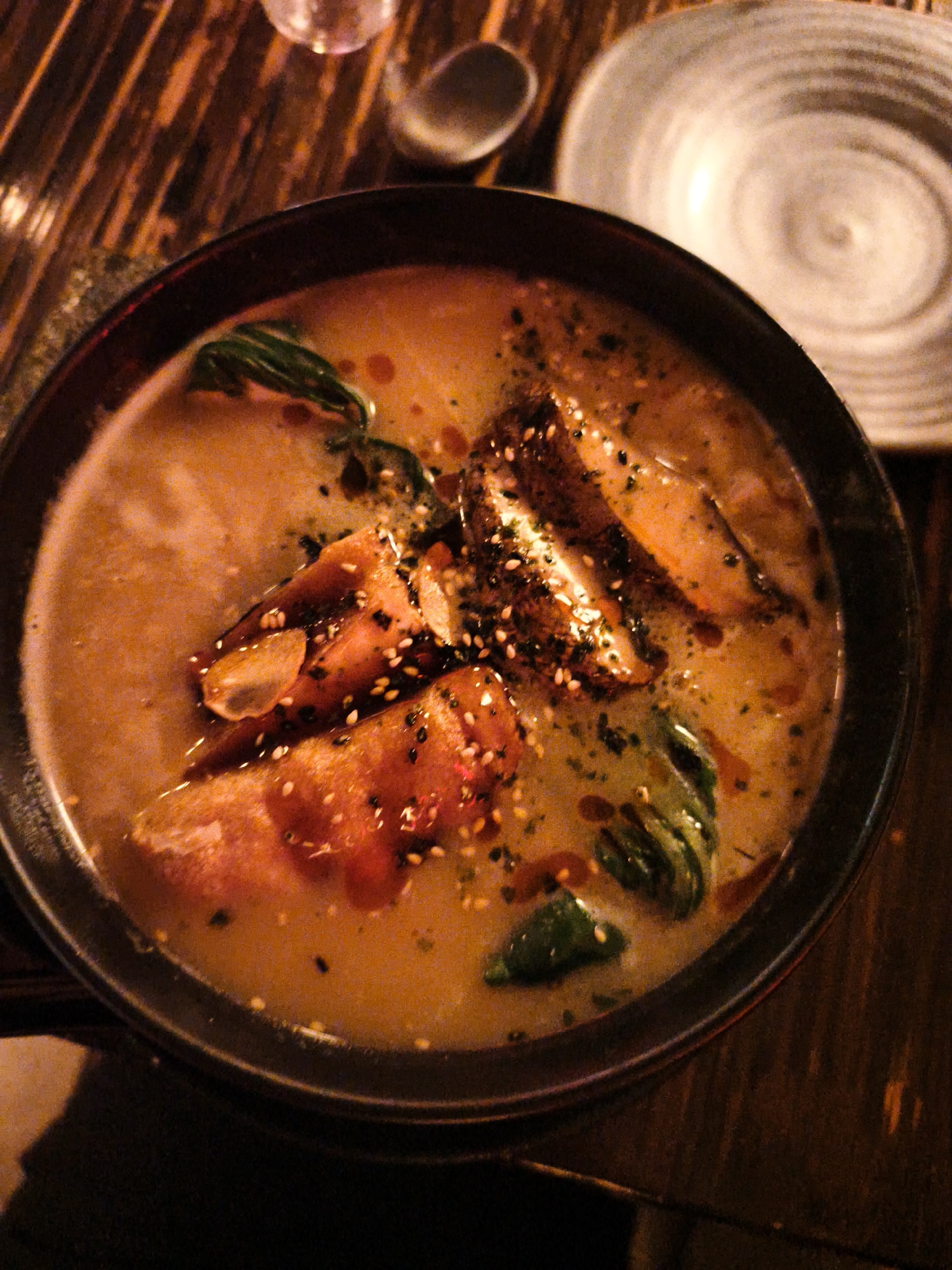 A close-up overhead shot captures a steaming bowl of ramen, brimming with a rich, orange broth. Slices of tender meat, possibly pork belly, are visible, some with a crispy, seared exterior, sprinkled with sesame seeds and a dark spice blend. Green vegetables, likely bok choy, add a splash of color and freshness to the dish. The ramen is served in a dark, round bowl with a textured rim, placed on a dark wooden table with a prominent grain. To the right of the bowl, a light-colored, patterned ceramic plate is partially visible, along with a small, grey utensil. The overall ambiance suggests a cozy, dimly lit restaurant setting. The image does not provide specific information about the location within Be'er Sheva, Israel, or the presence of any individuals.